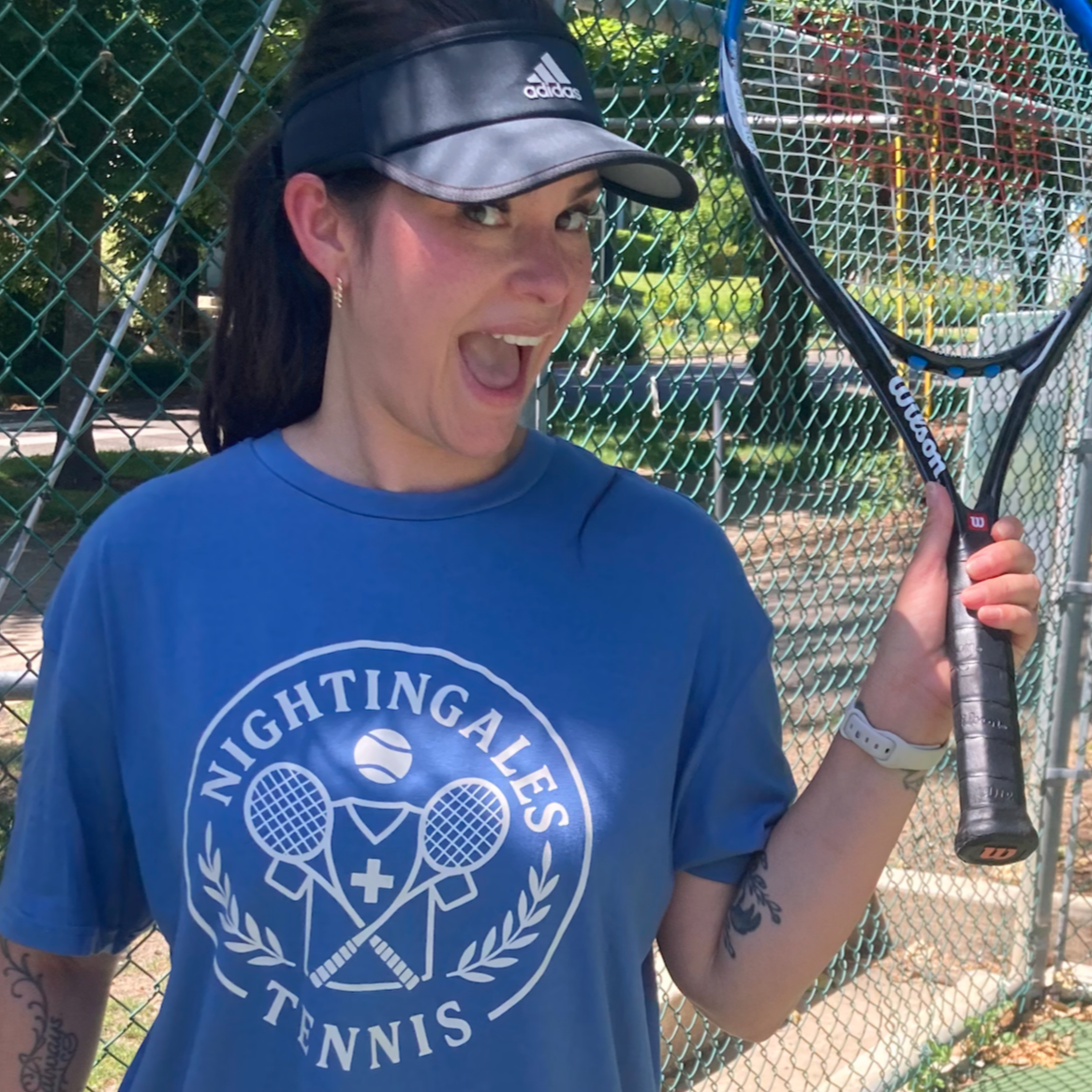 Woman wearing a black Adidas visor, blue Nightingales Tennis T-shirt, holding a Wilson tennis racket, standing in front of a chain-link fence at an outdoor tennis court, smiling and looking excited.