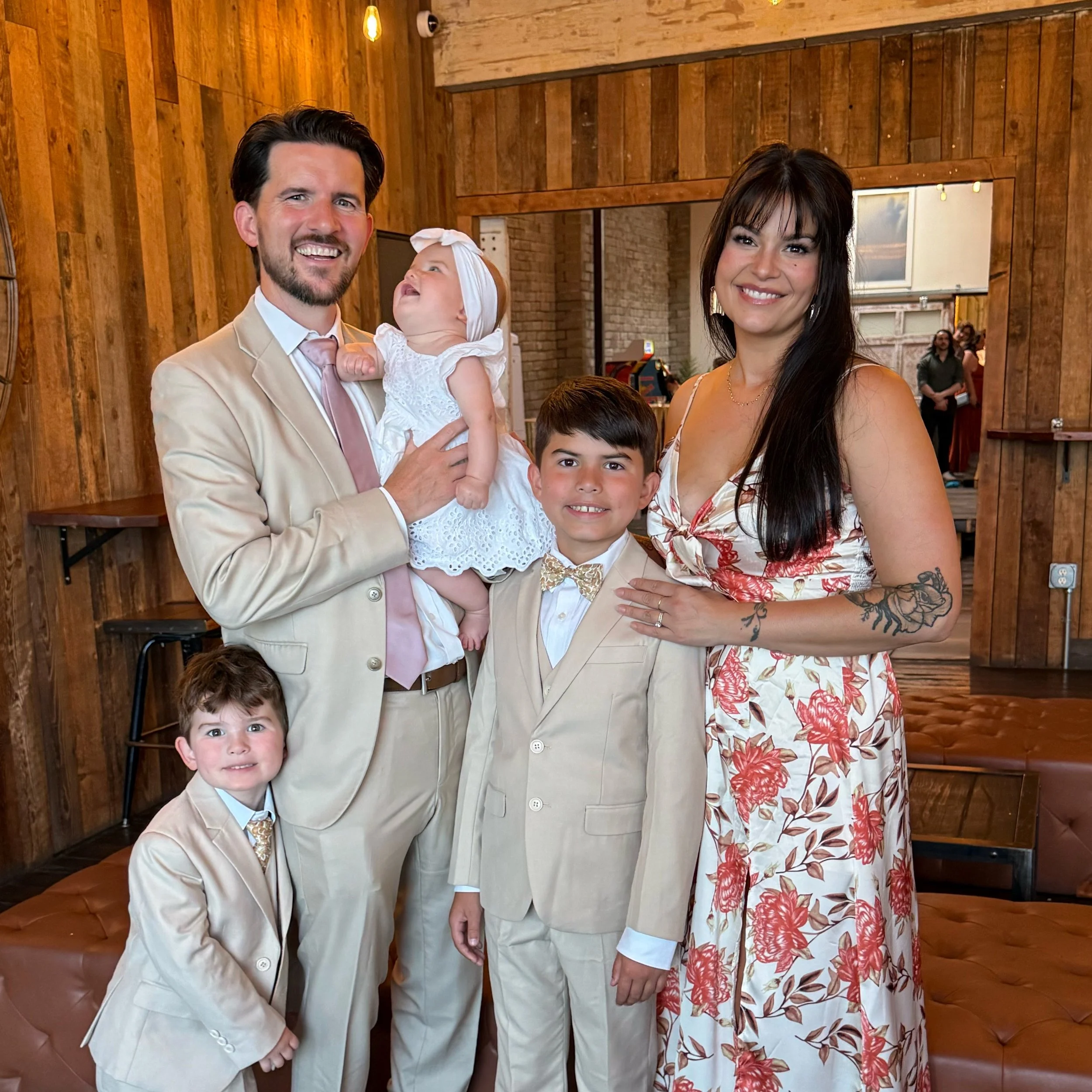A family of five posing indoors with wooden wall background. The father in a beige suit holding a baby girl in white, mother in a floral dress standing next to their two young sons in matching suits.
