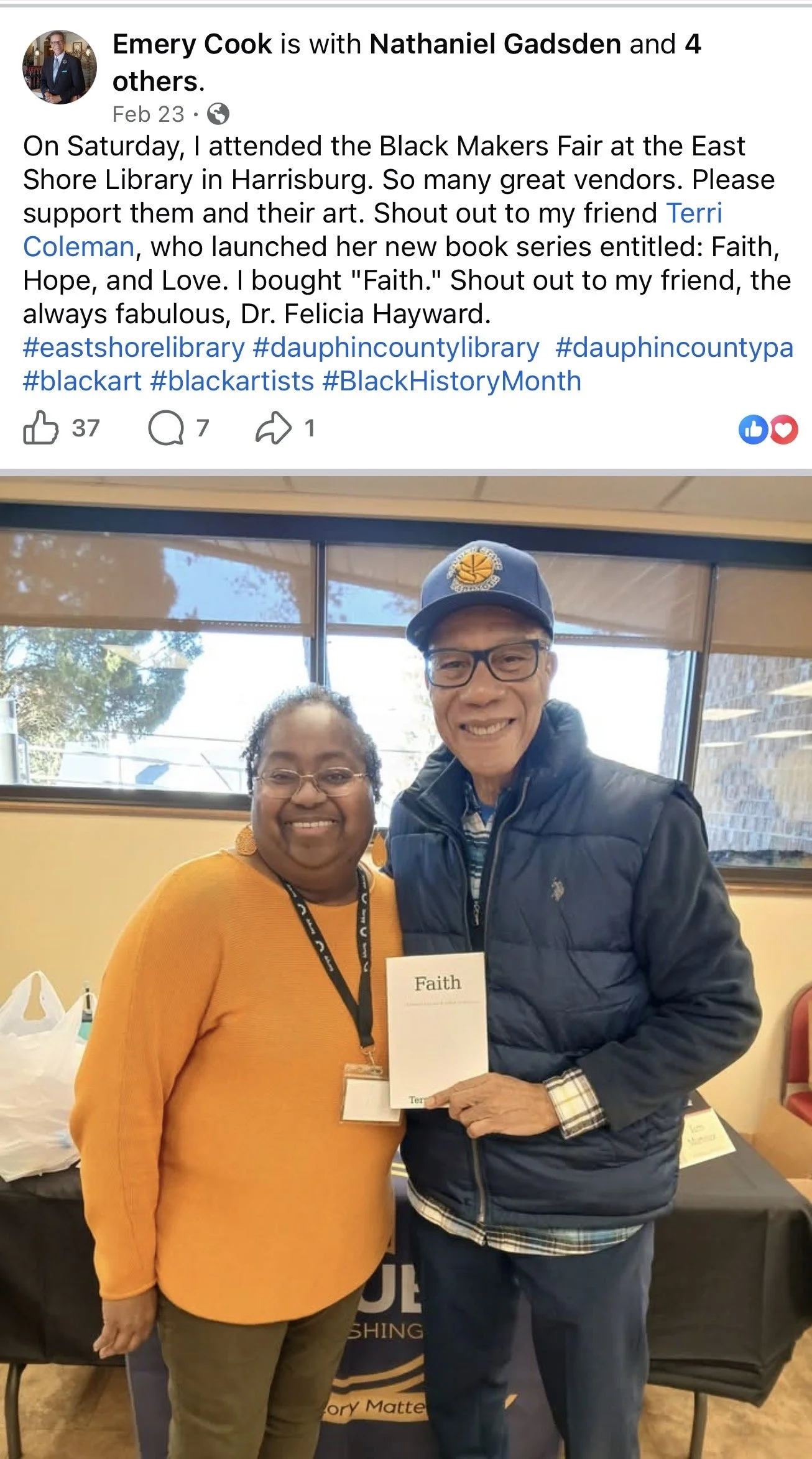 Two people, a woman in an orange shirt and a man in a navy jacket, standing indoors and smiling at the camera. The woman is wearing glasses, earrings, and a name badge. The man is wearing a baseball cap, glasses, and is holding a small card with the 
