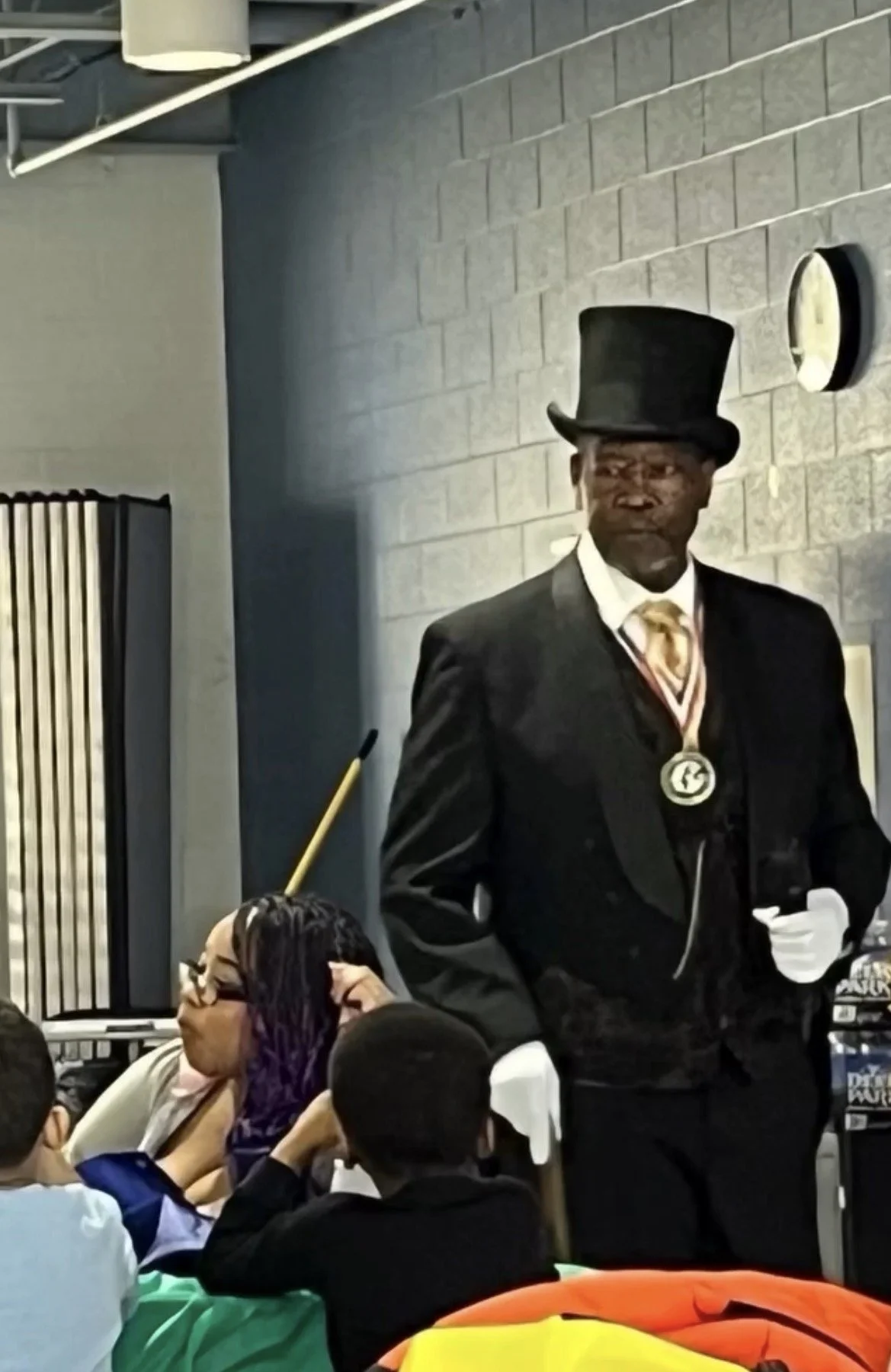A man dressed in vintage formal attire, including a black suit, a striped tie, a white shirt, white gloves, and a tall black top hat, standing indoors near a crowd of people. The setting appears to be an event or gathering.