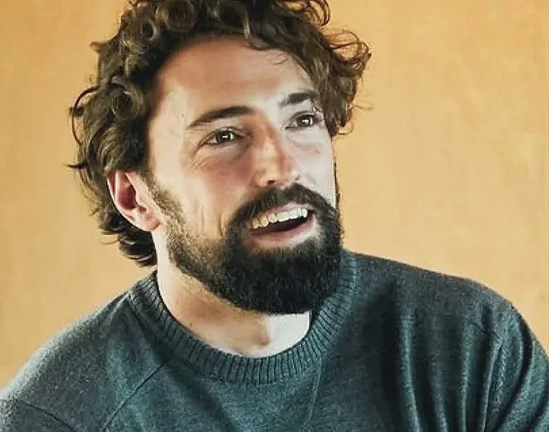 A young man with curly brown hair and a full beard, smiling and looking to the side, wearing a dark gray sweater, against a plain light brown background.
