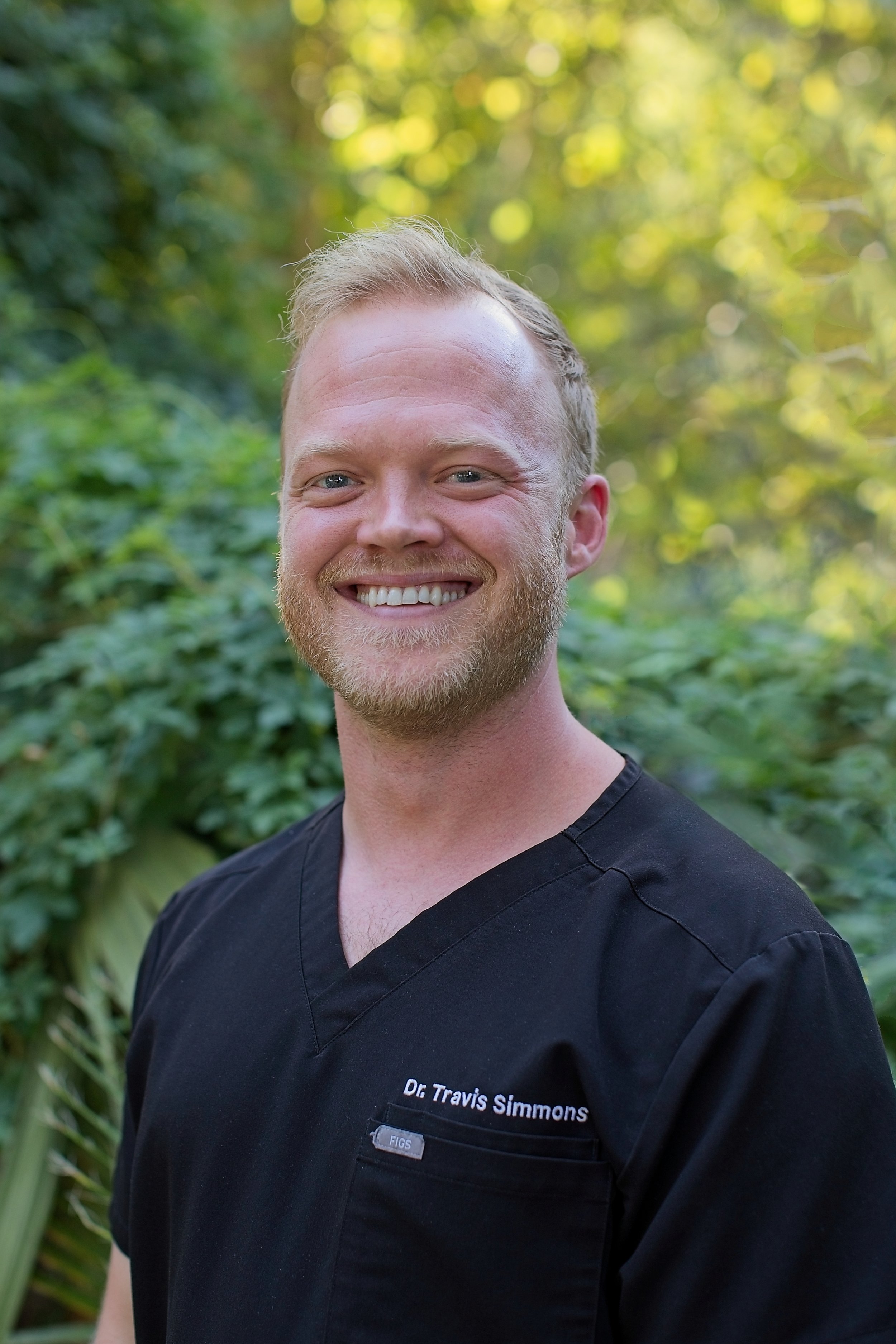 A kind male doctor with a beard and short blond hair smiling outdoors in front of green foliage, wearing a black medical scrub top with the text 'Dr. Travis Simmons' embroidered on it. Primary care. Direct Primary Care. New Braunfels.