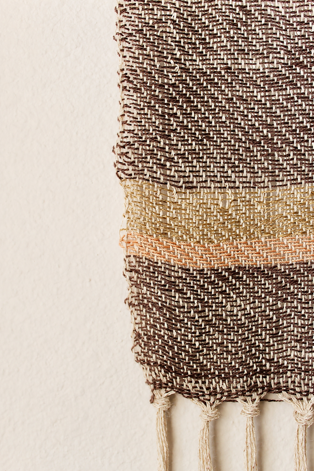 Close-up of a woven wall hanging with horizontal stripes in brown, beige, peach, and black, with tassels hanging from the bottom.