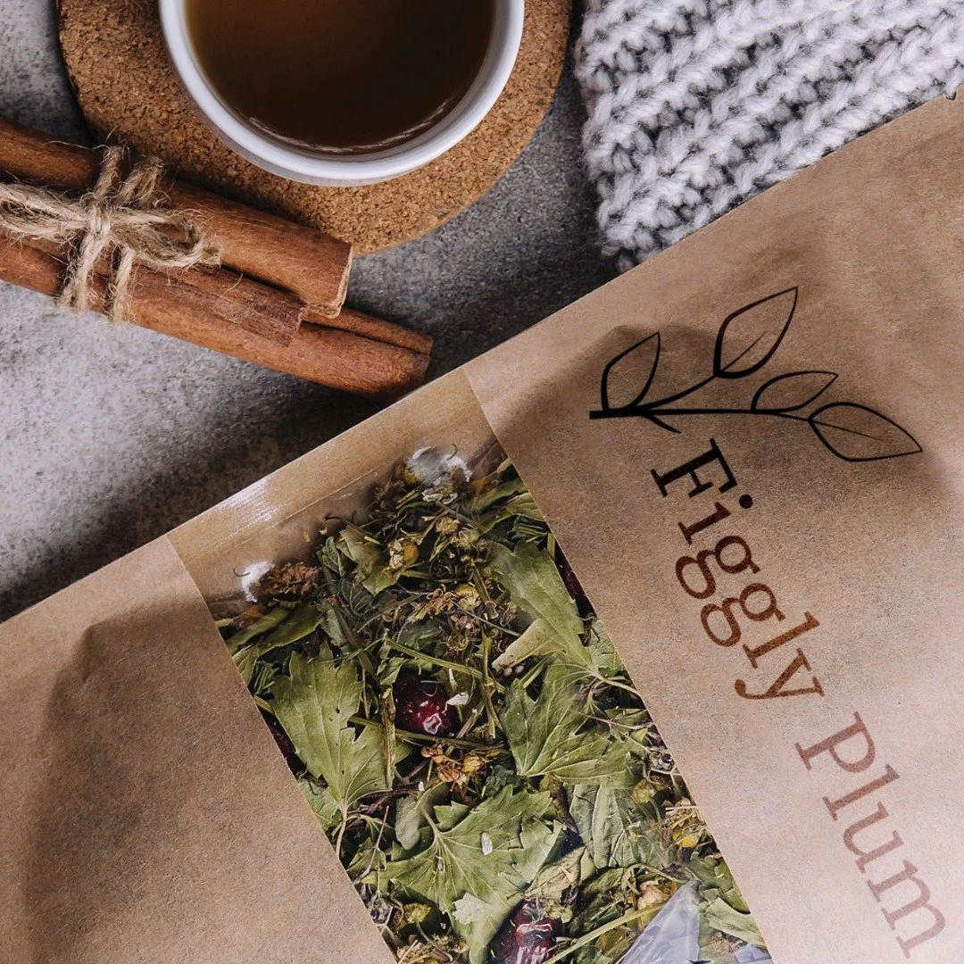 A paper pouch labeled 'Eco-friendly Plum' with a leaf design, containing dried mixed herbs or leaves. Next to it is a cup of tea on a cork coaster, a bundle of cinnamon sticks tied with twine, and a gray and white knitted cloth.