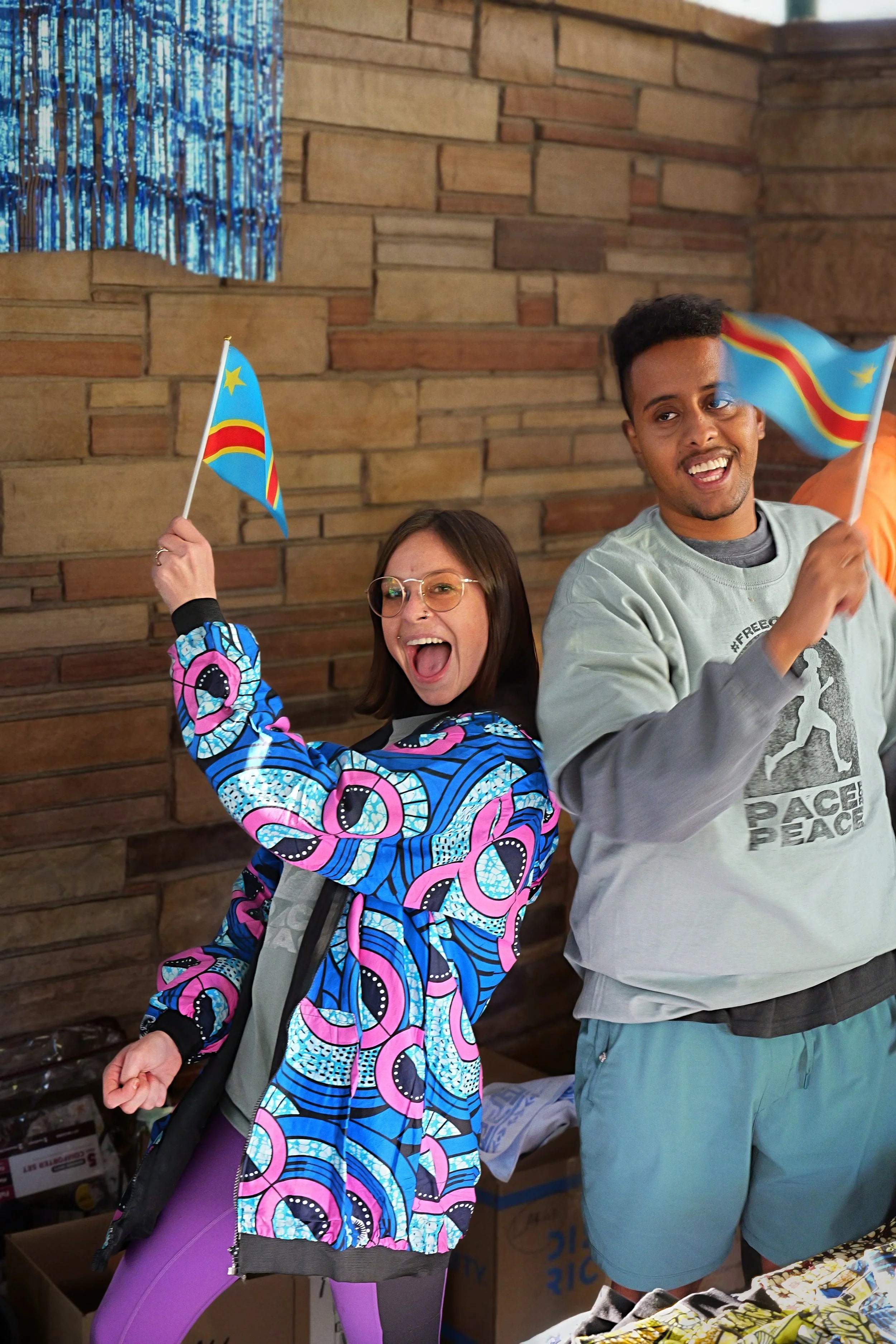 Two smiling people celebrating with small Congolese flags, standing indoors against a brick wall. One person wears glasses and a vibrant patterned jacket, and the other is dressed in a light gray sweatshirt and shorts.