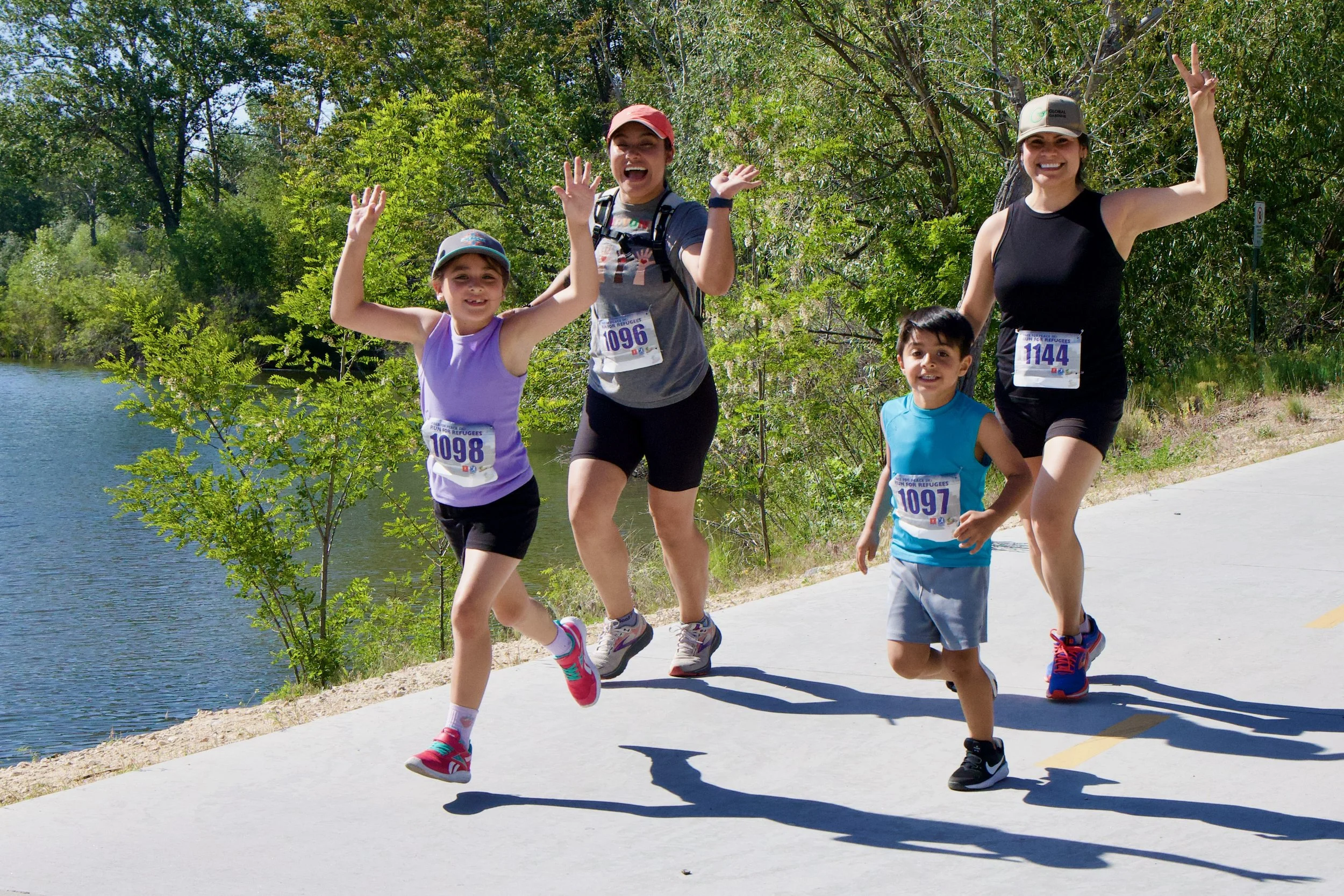 Four people, two adults and two children, participating in a fun run by a lake, smiling and making peace signs. All are wearing bib numbers and athletic clothing, with trees and water in the background on a sunny day.