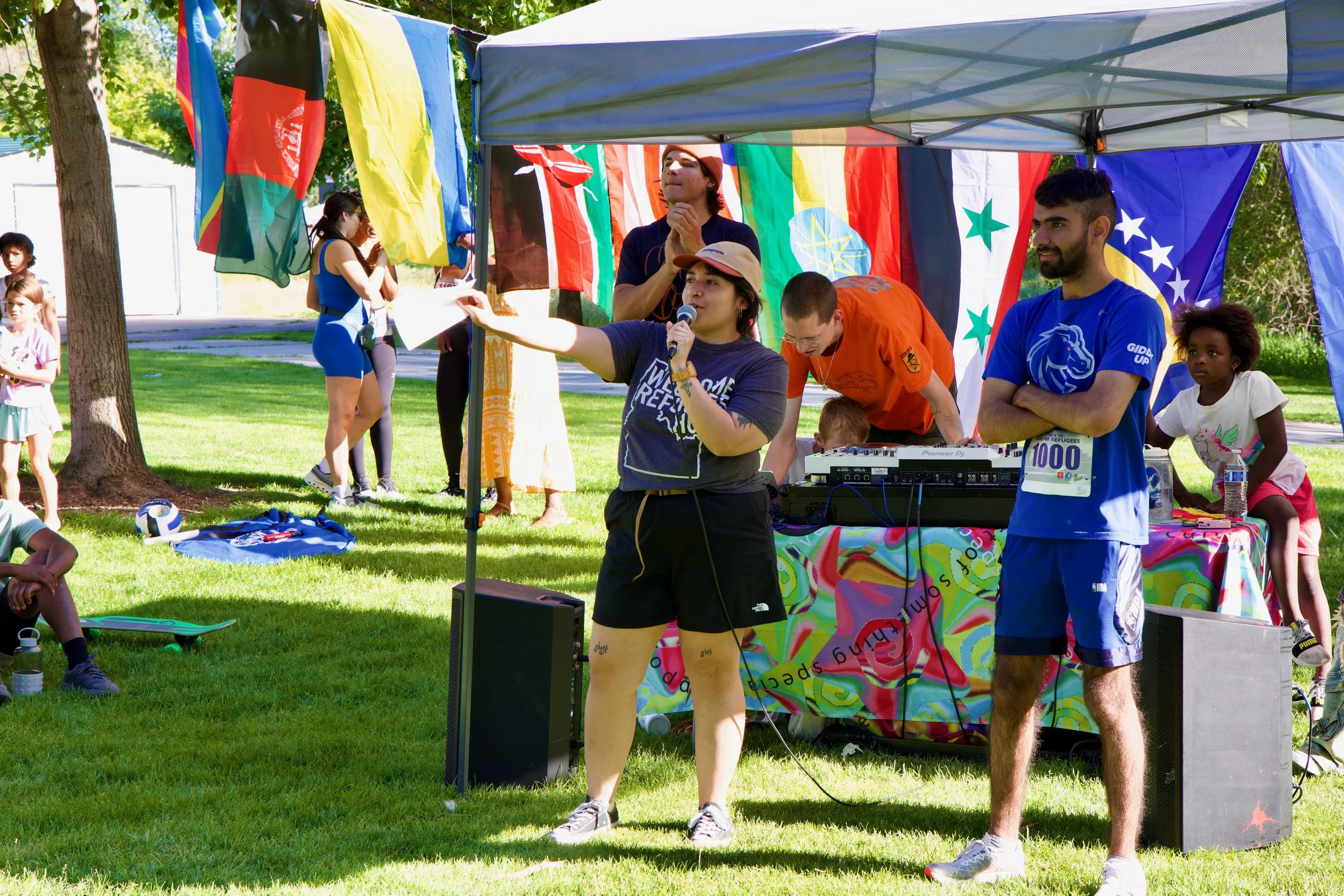 People gathered outdoors under a tent decorated with international flags, with a woman speaking into a microphone and a man standing next to DJ equipment, at a multicultural community event.