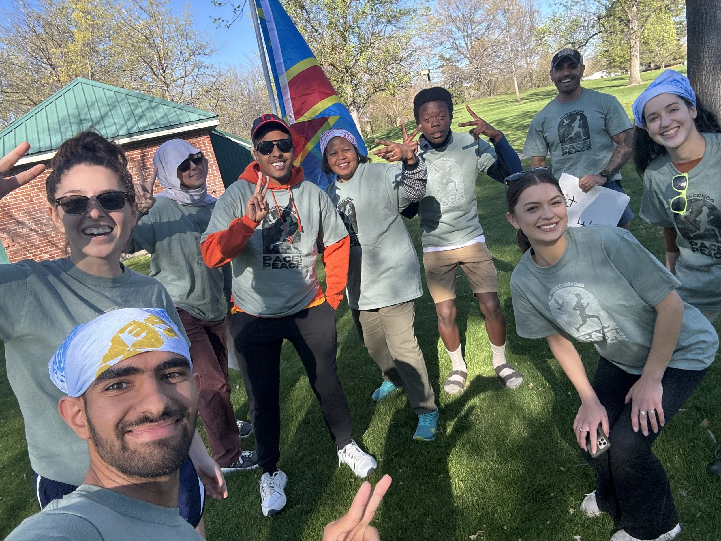 Group of people outdoors, smiling, making peace signs, participating in an event with a colorful flag, on a sunny day with trees and grass.