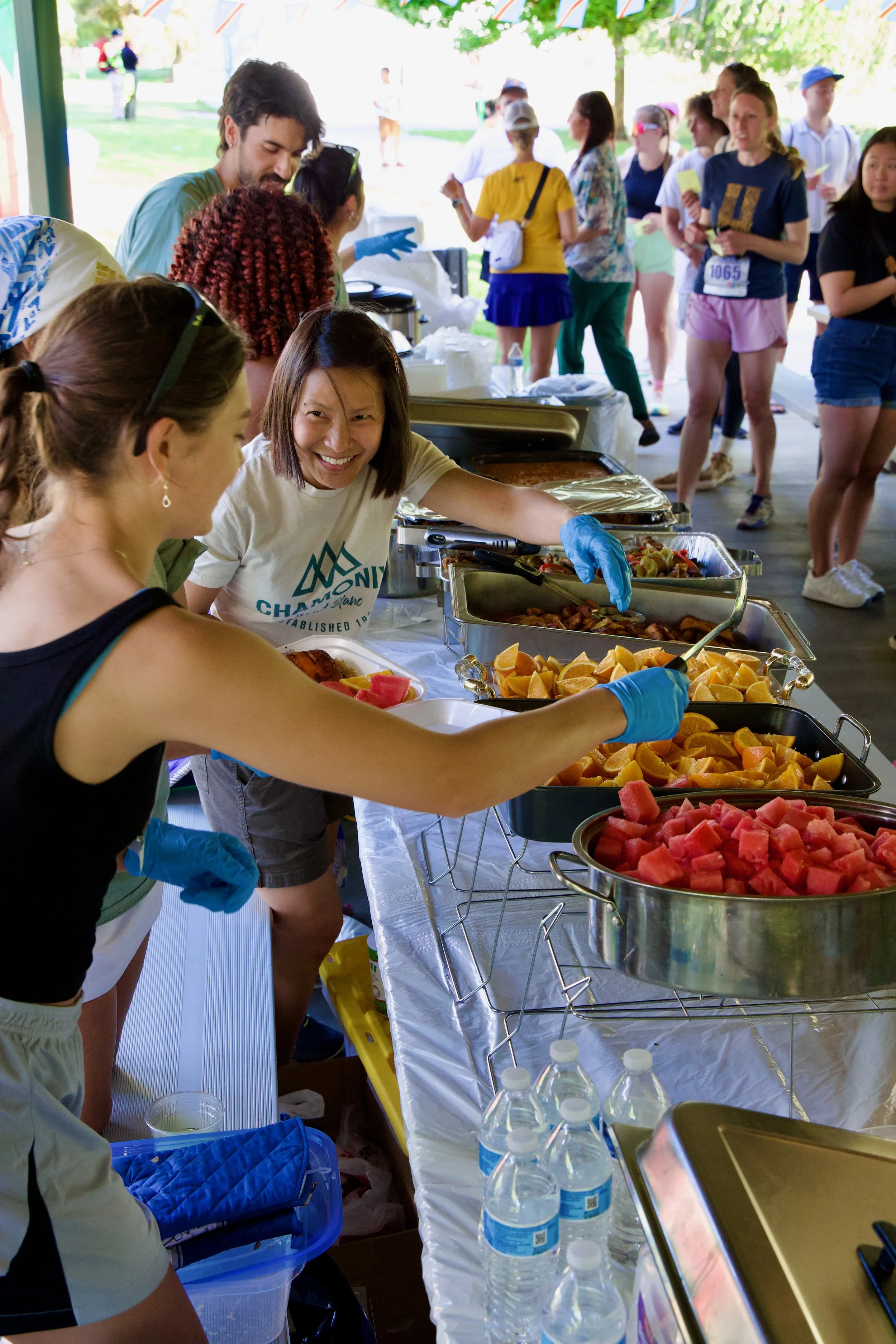 People serving and collecting food at a buffet table with trays of sliced fruits such as watermelon, oranges, and others, at an outdoor event.