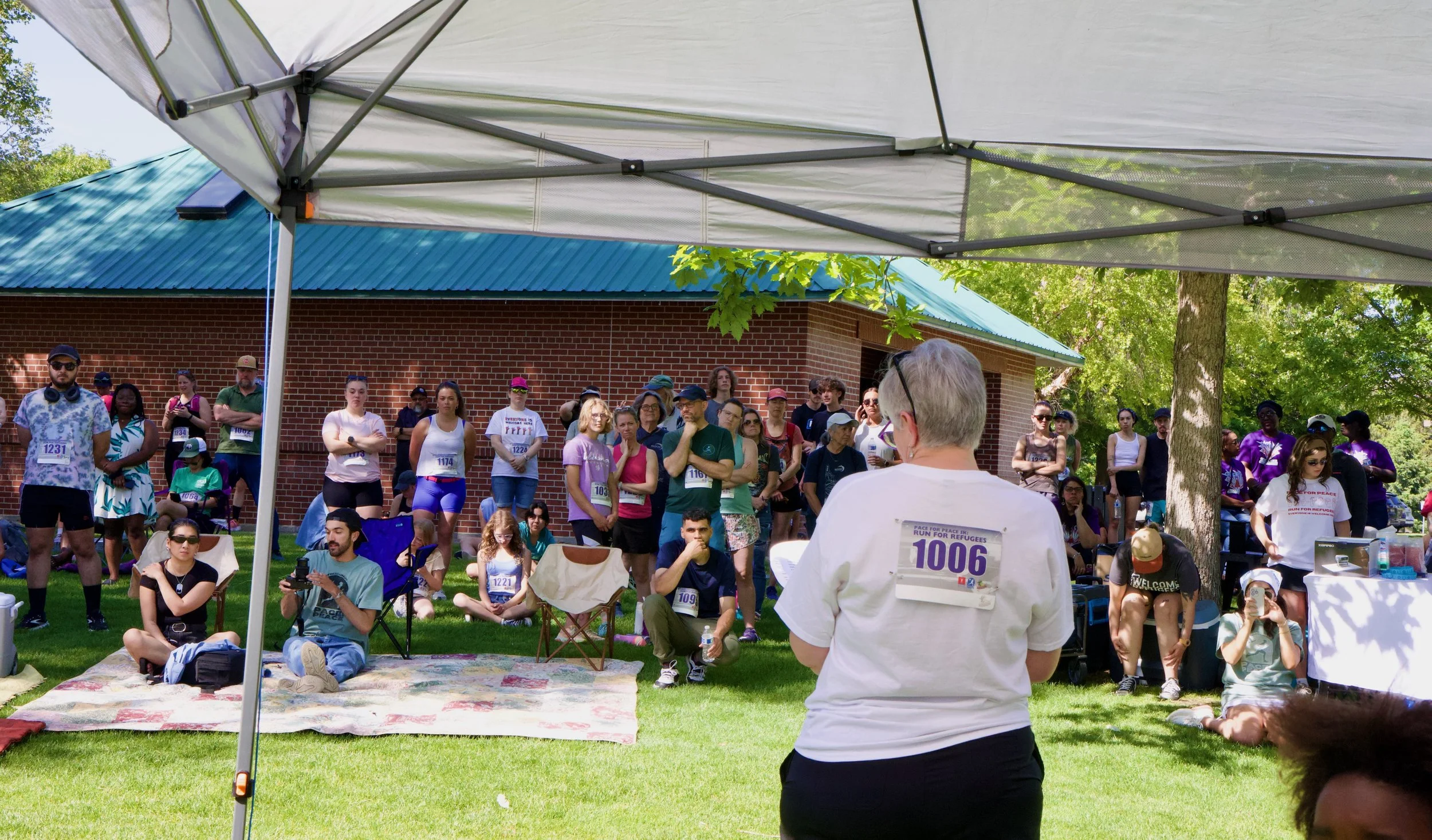 A group of diverse people gathered outdoors under a tent, listening to a woman with short gray hair speaking. Many attendees are seated or standing, some with race bibs, in front of a brick building with a blue roof, surrounded by green trees.