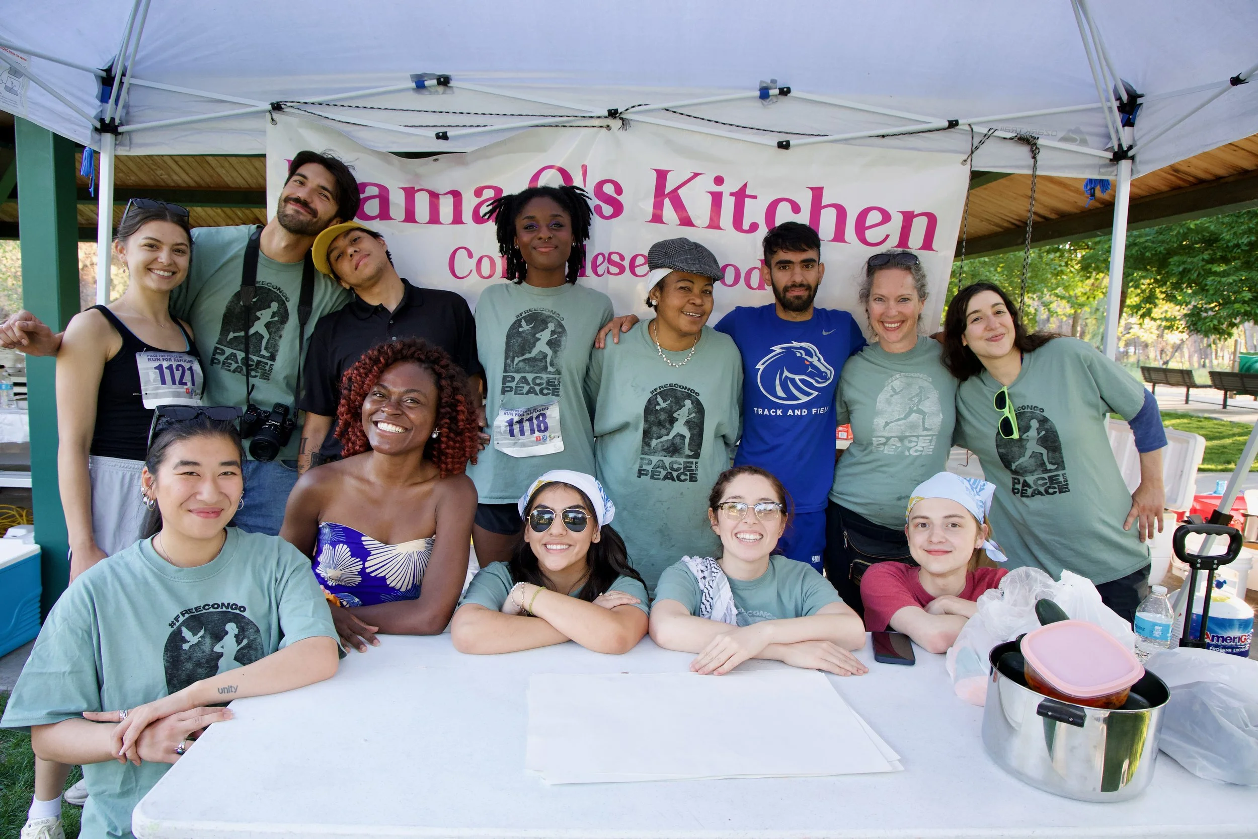 Group of diverse people smiling and posing at a community event under a tent with a banner that says "Mama Cs Kitchen" and features children running, some seated at a table with food, in an outdoor park.