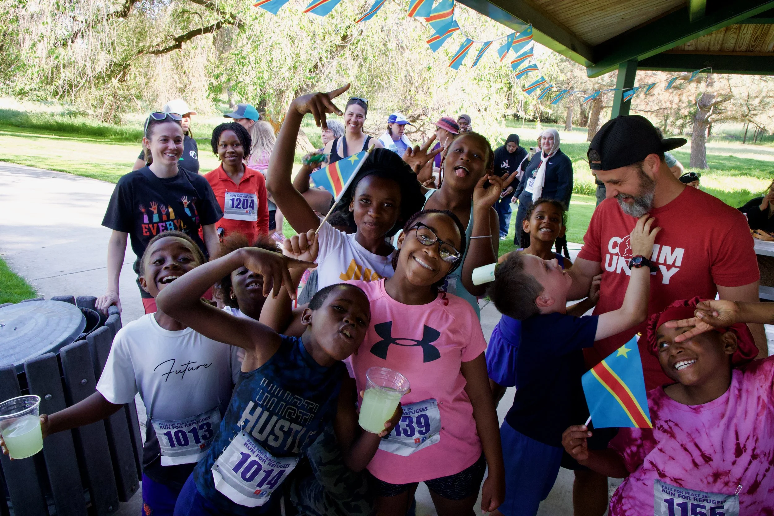 Group of children and a few adults at an outdoor event, some holding small flags, smiling and making peace signs, with a park and trees in the background.