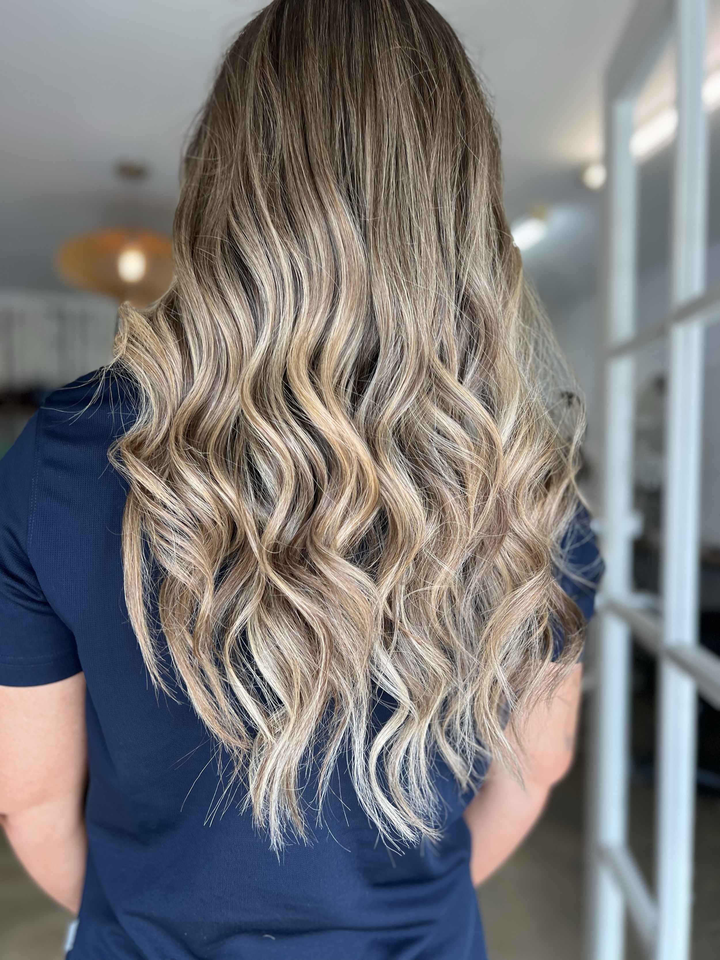 Back view of a woman with long, wavy blonde hair wearing a navy blue shirt indoors.