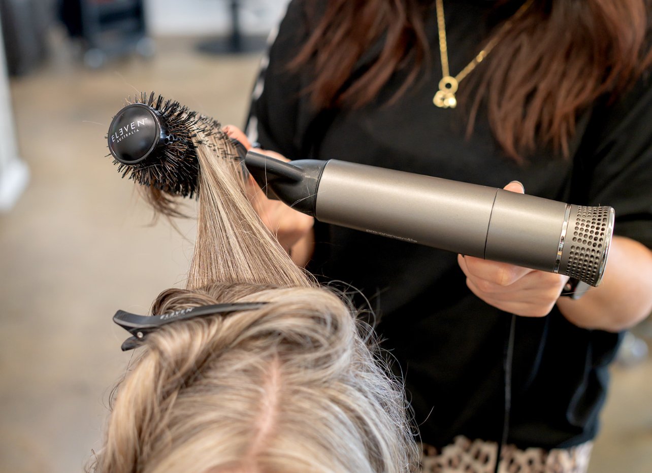 A hairstylist drying a client's hair with a blow dryer and round brush in a salon.