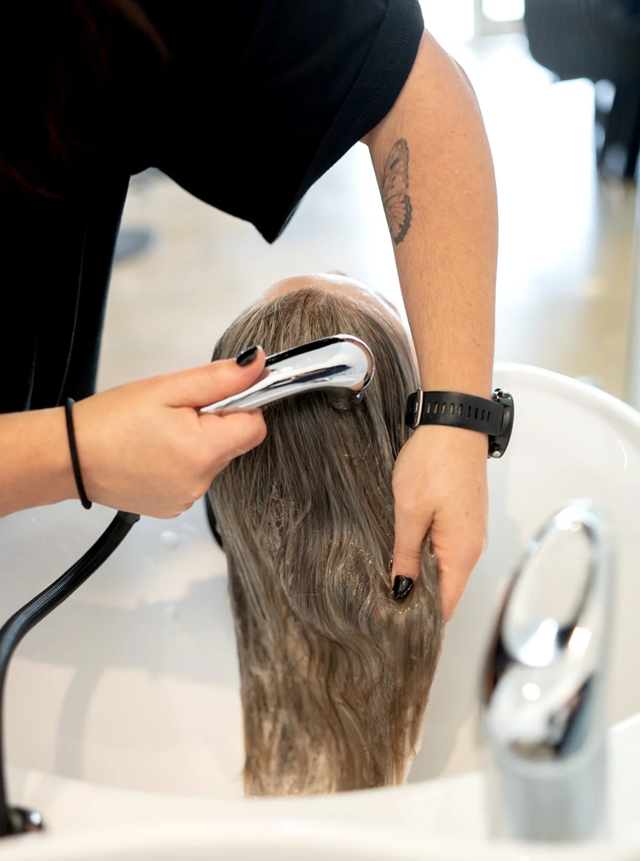 A person with long hair receiving a hair treatment or shampoo at a salon, with a hairstylist using a handheld device on their hair over a white sink.