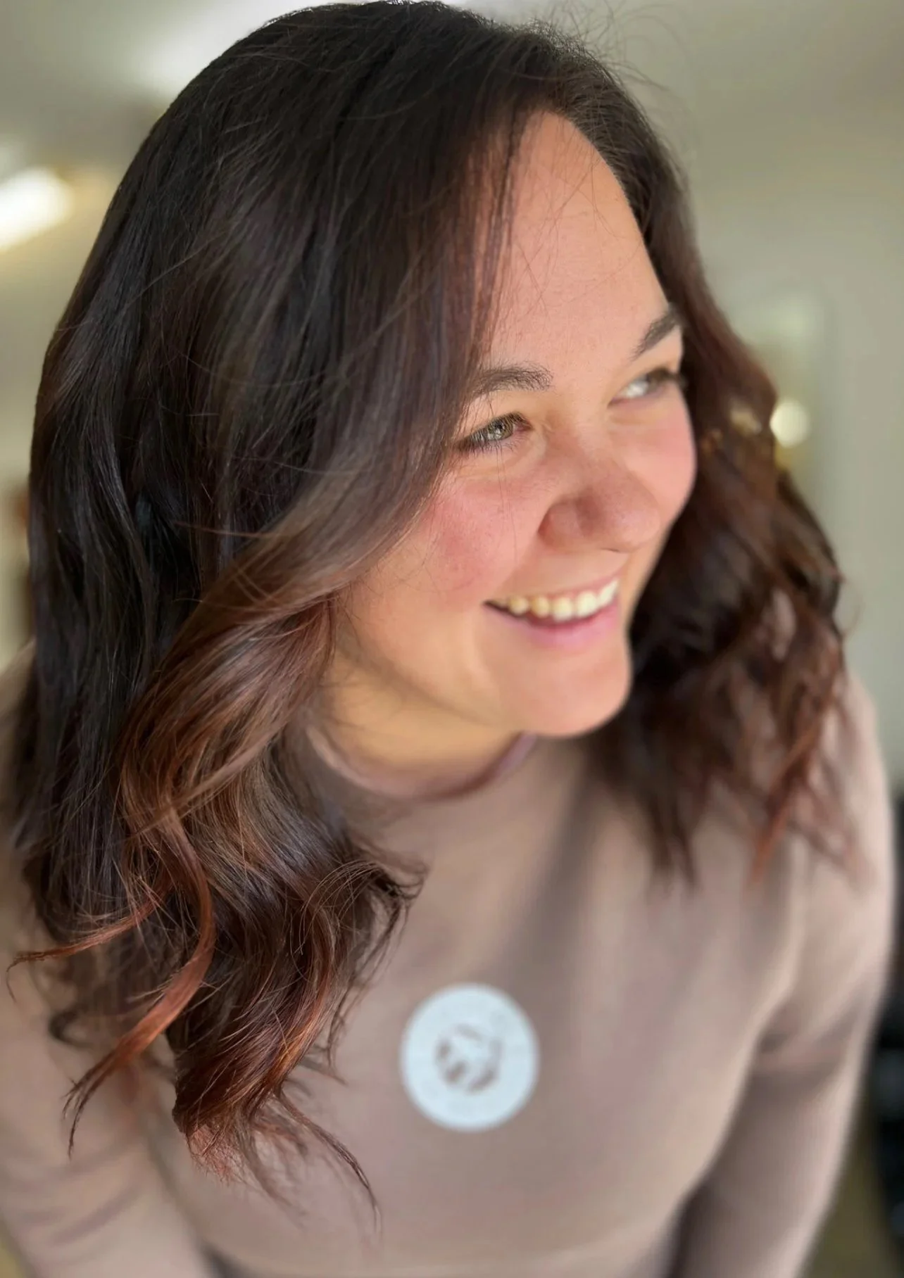 A smiling woman with wavy brown hair looking to her left, wearing a beige top with a white circular sticker on it.