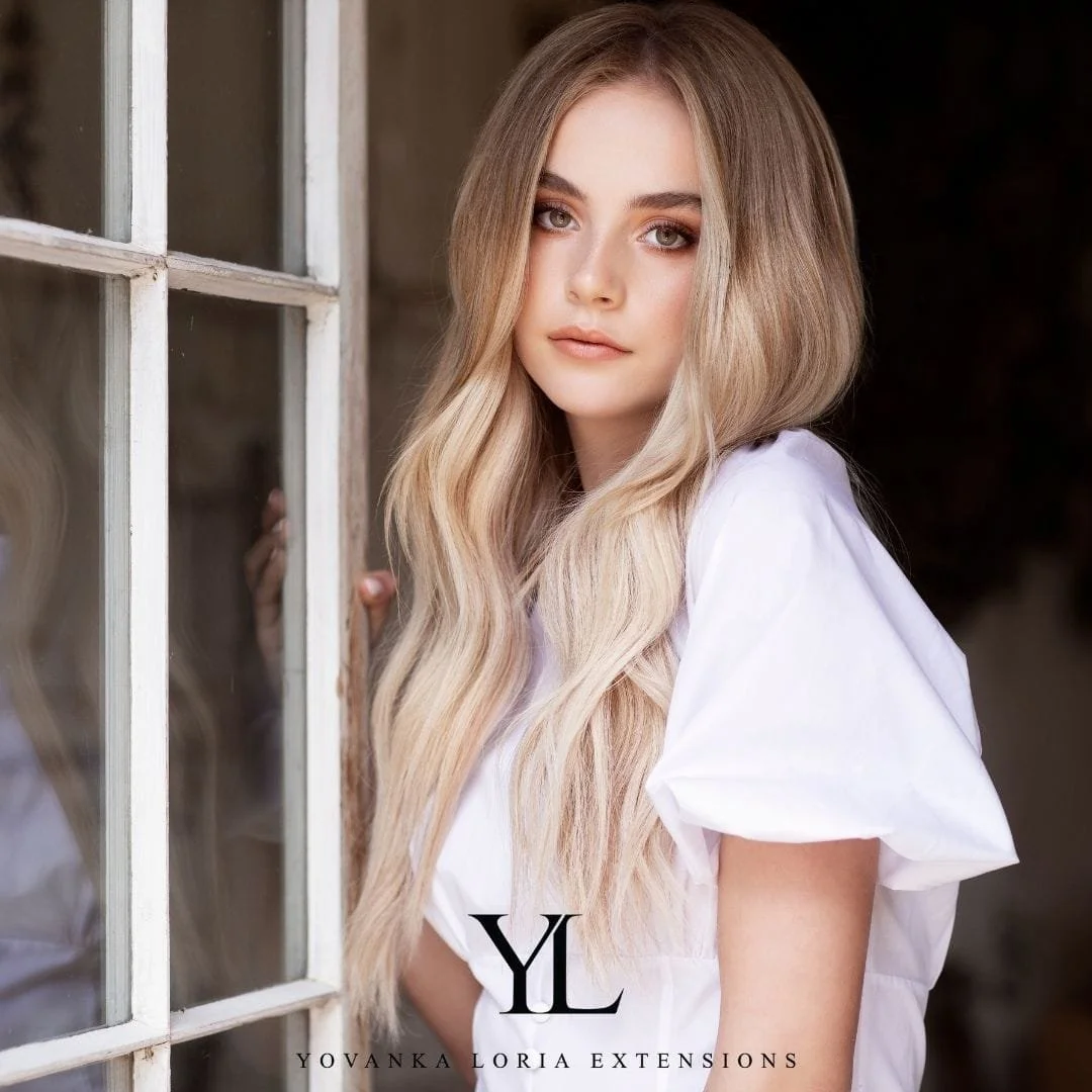 A young woman with long, wavy blonde hair standing next to a window with white framing, wearing a white top, looking at the camera. weft hair extension