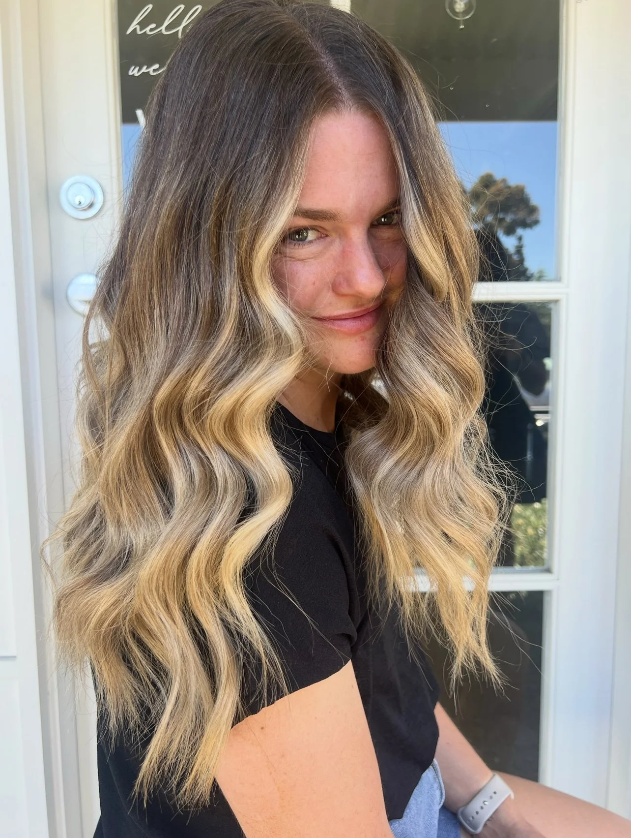 A woman with long, wavy blonde hair, sitting outdoors near a white door with window panes. She is wearing a black shirt and a smartwatch on her left wrist, with a subtle smile and a slightly mischievous expression.