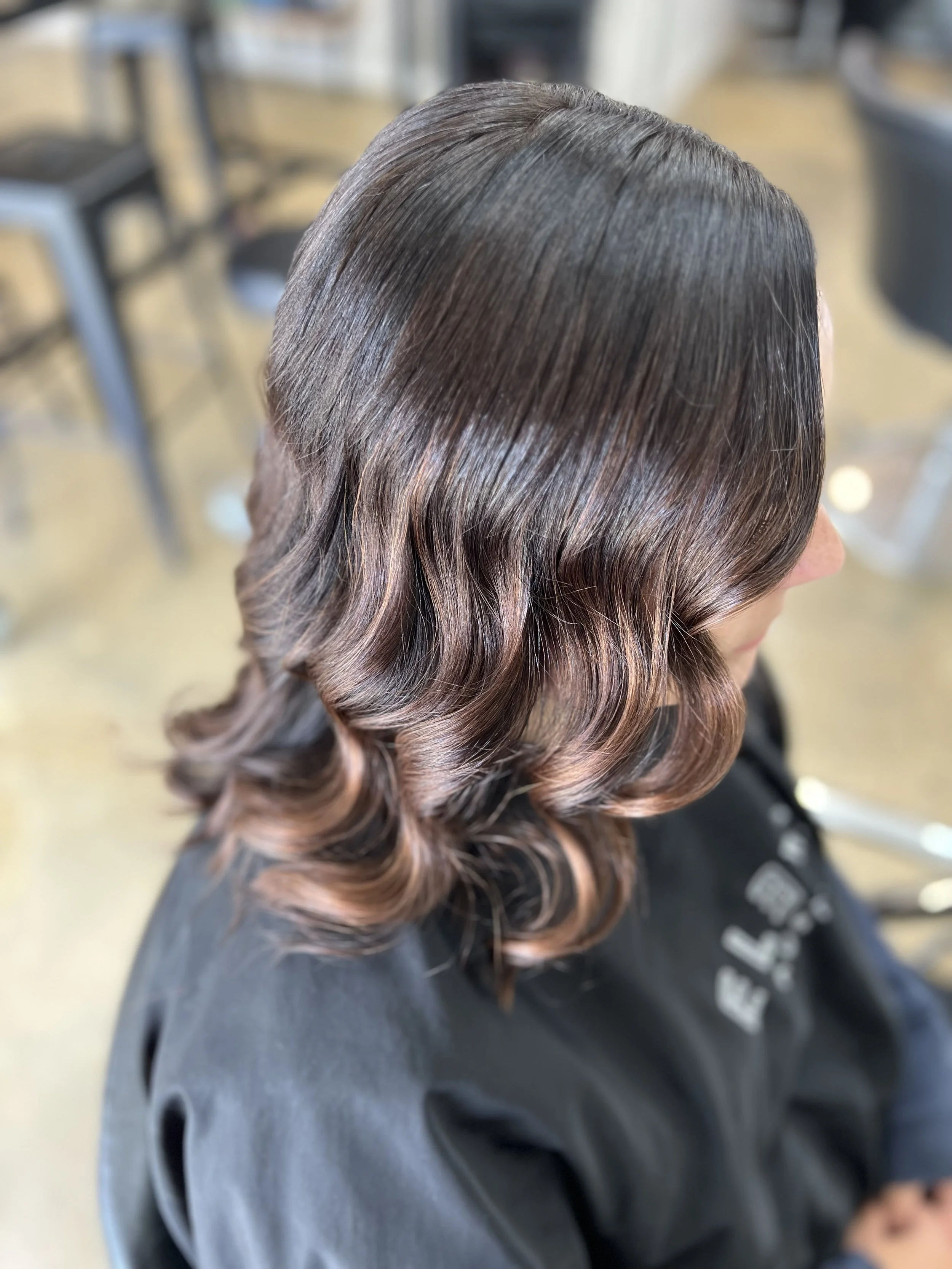 A woman's hair styled in soft, loose curls with a shiny, smooth finish and balayage highlights from dark brown to copper.