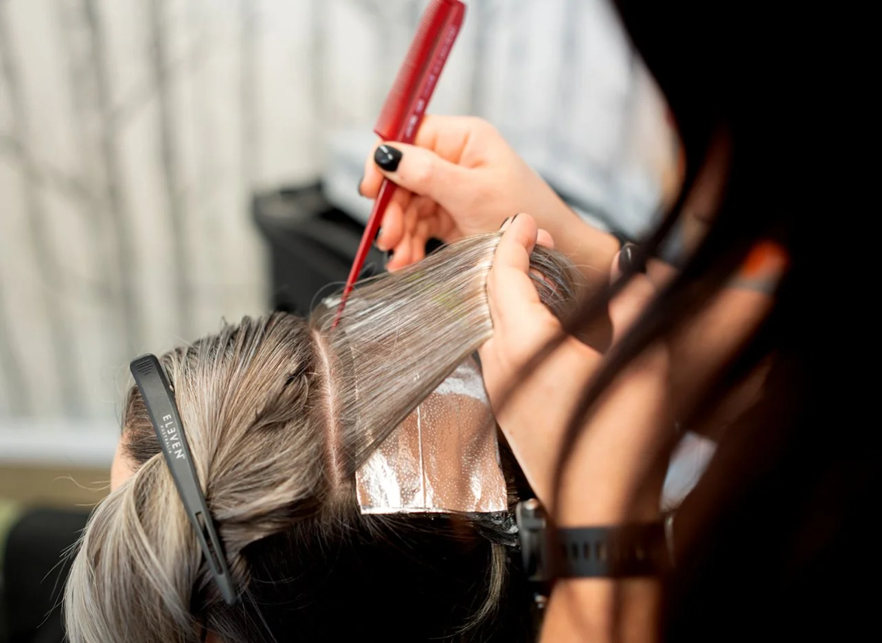 A hairdresser applies hair color to a client's hair using a thin brush, with the client wearing a protective cape and hair clips, in what appears to be a salon.