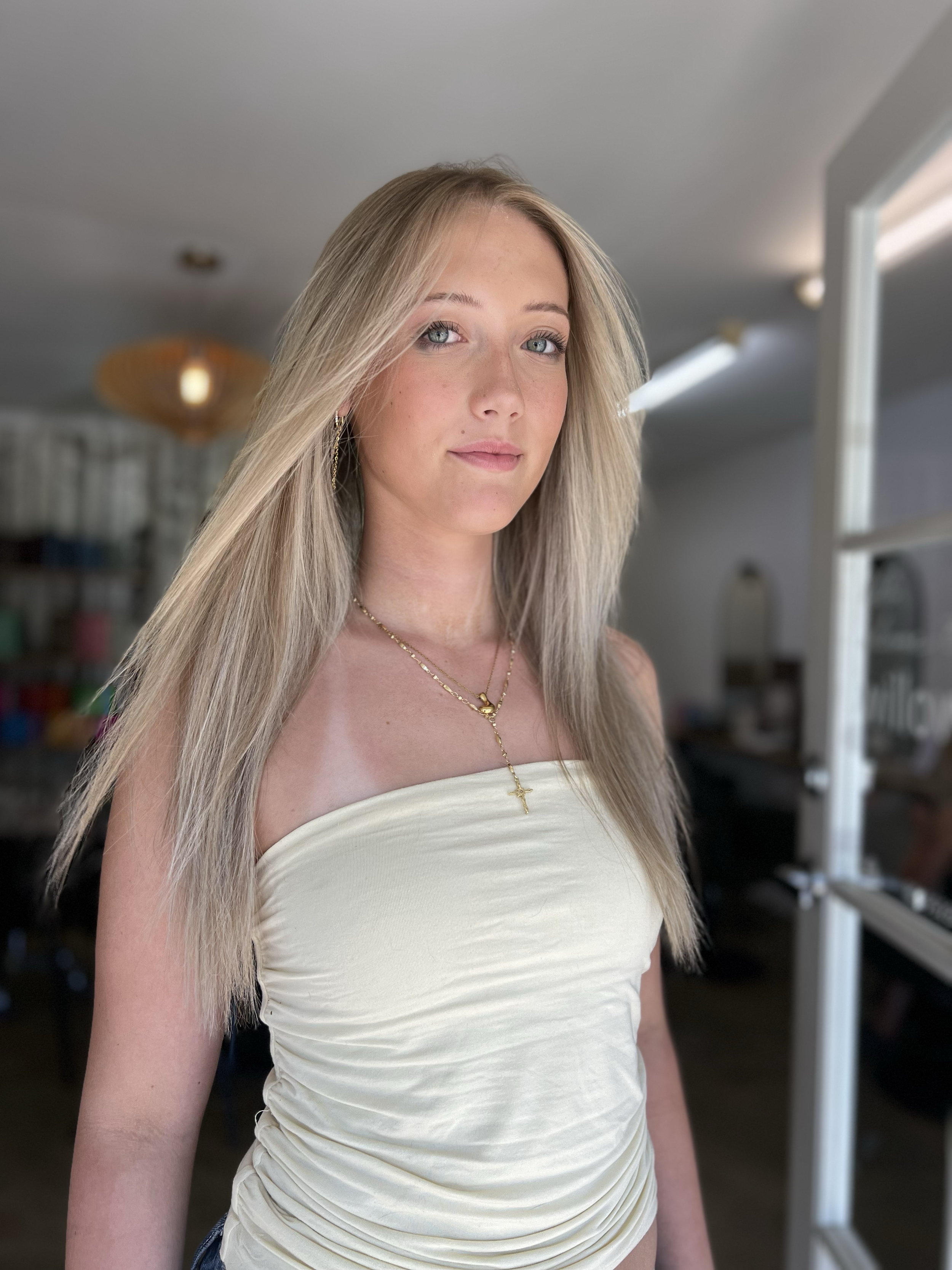 A young woman with long blonde hair, blue eyes, and light skin wearing a strapless beige top and gold jewelry, standing indoors with a blurred background of a room.