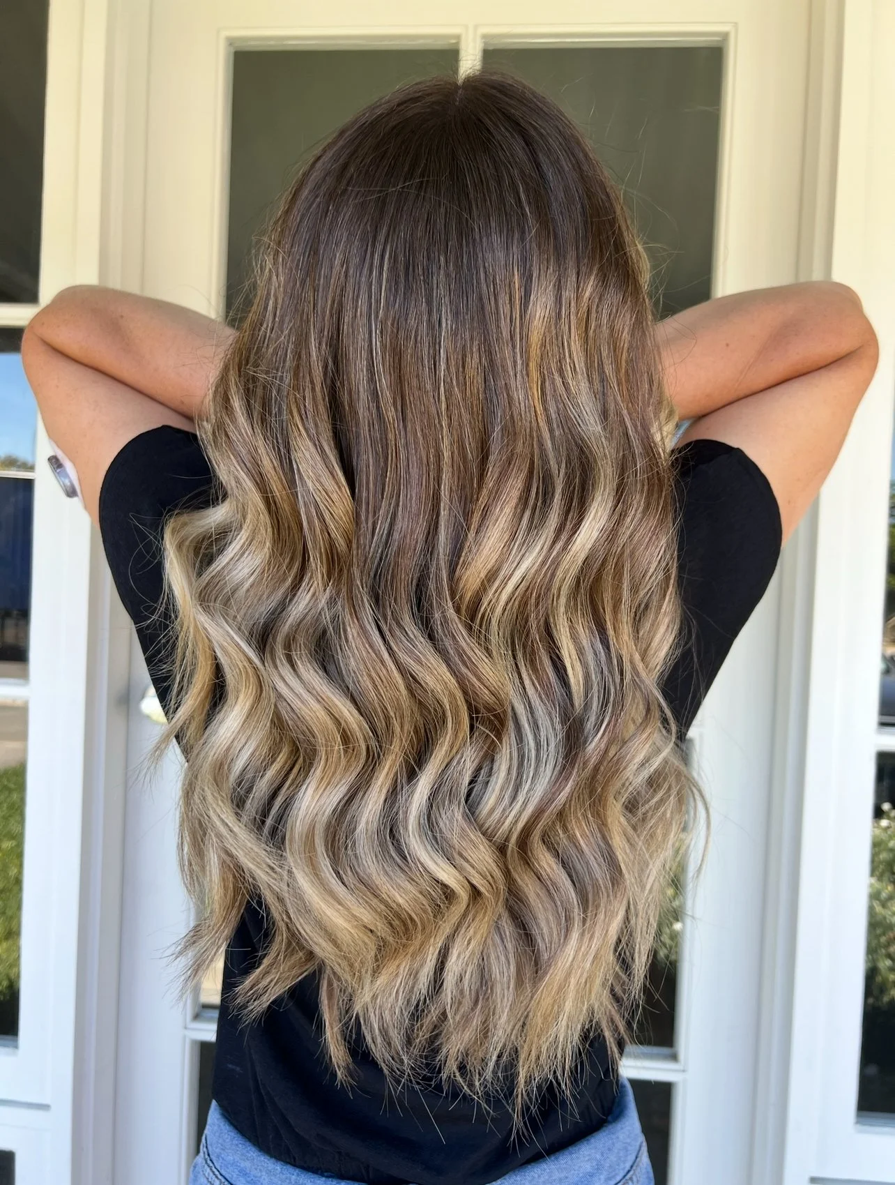 Back view of a woman with long wavy hair standing outside in front of a white door, wearing a black shirt and blue jeans.