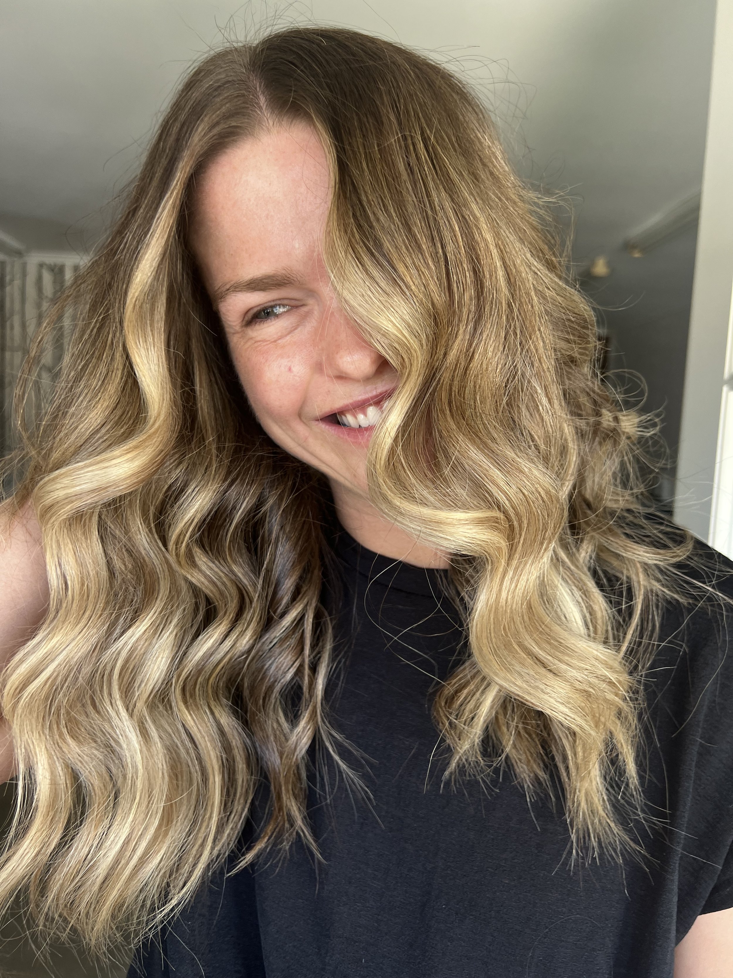 A woman with long wavy blonde hair and a black shirt, smiling and looking to her right, in a bright room with neutral-colored walls.