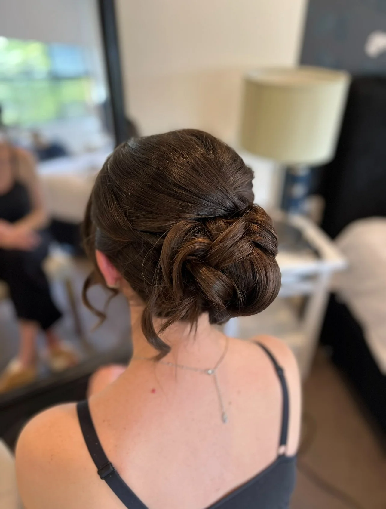 Back of a woman with dark brown hair styled in an elegant updo with braids and curls, wearing a black strap top and a delicate necklace, sitting in a room with a mirror, lamps, and blurred background.
