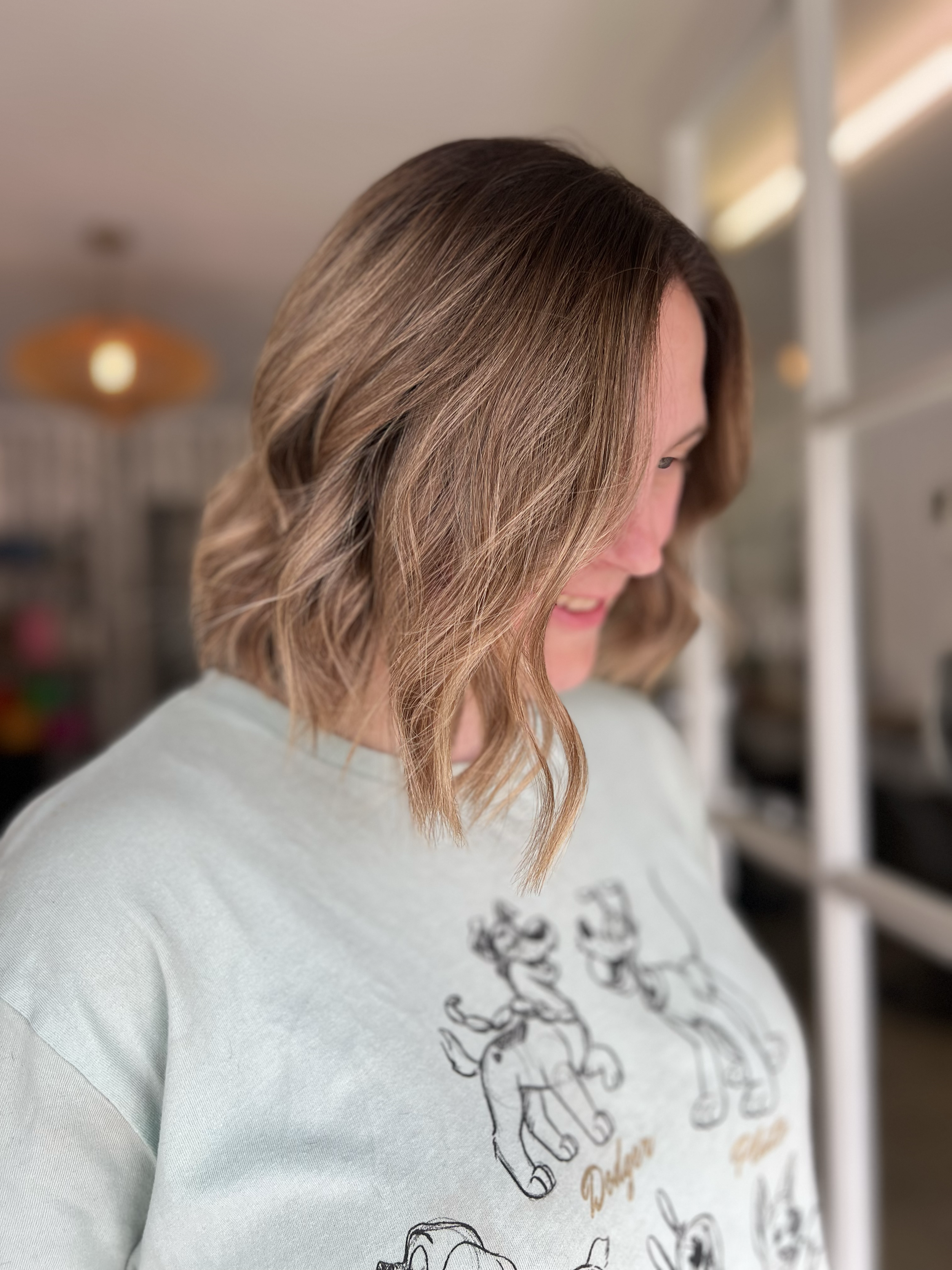 Side view of a woman with shoulder-length wavy brown hair, smiling and looking down, wearing a light-colored T-shirt with cartoon dog illustrations.