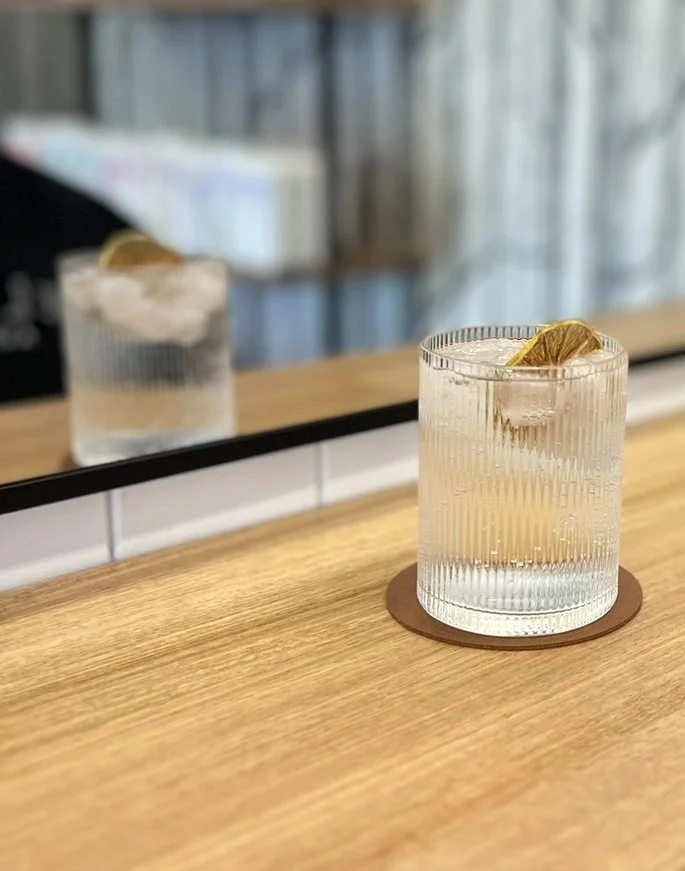 A clear glass with vertical ridges containing a lemon wedge, placed on a round coaster on a wooden surface, with a mirror reflecting the glass and a blurred background.