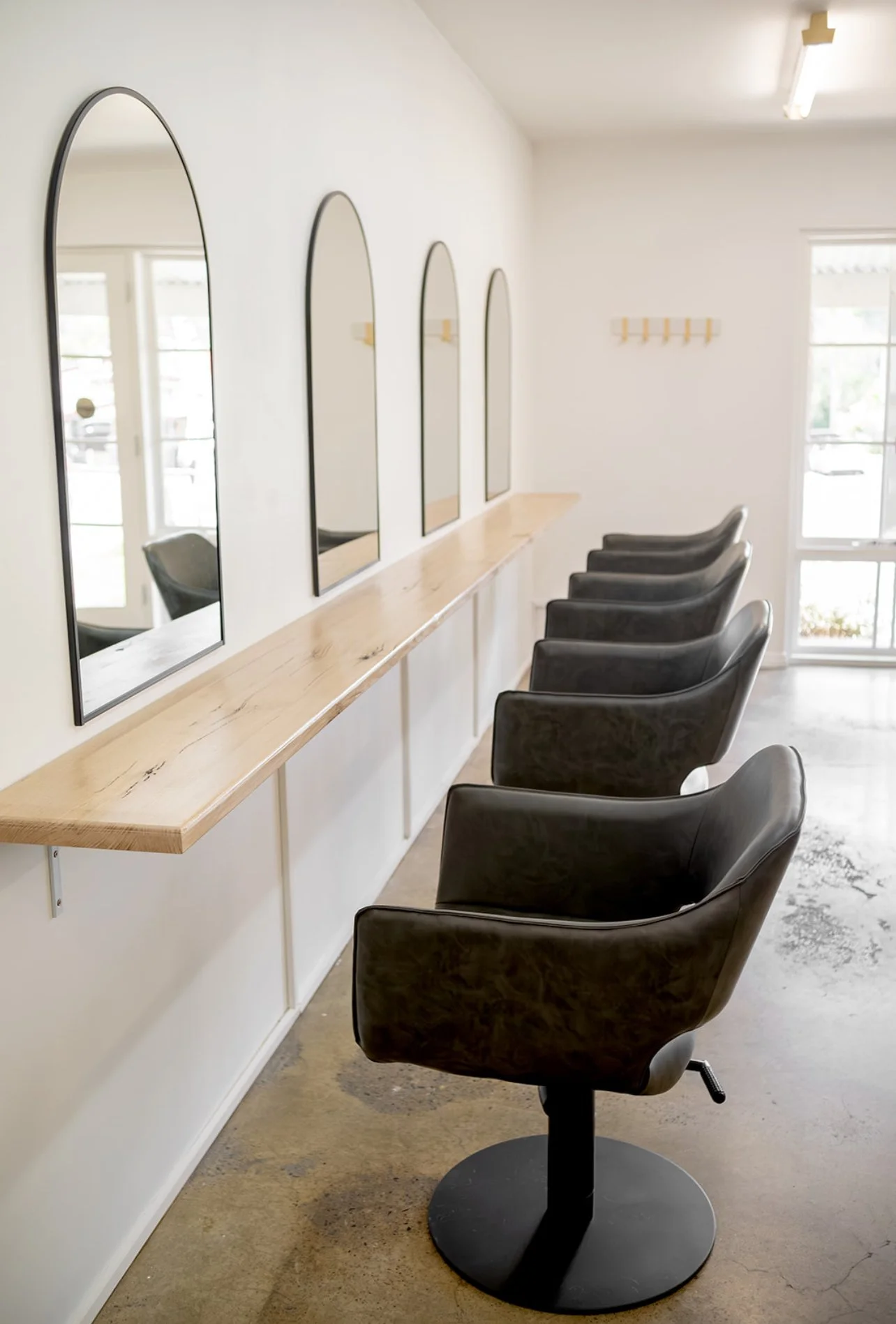 Empty hair salon with four black styling chairs in front of a long wooden mirror station with four curved mirrors on the wall.
