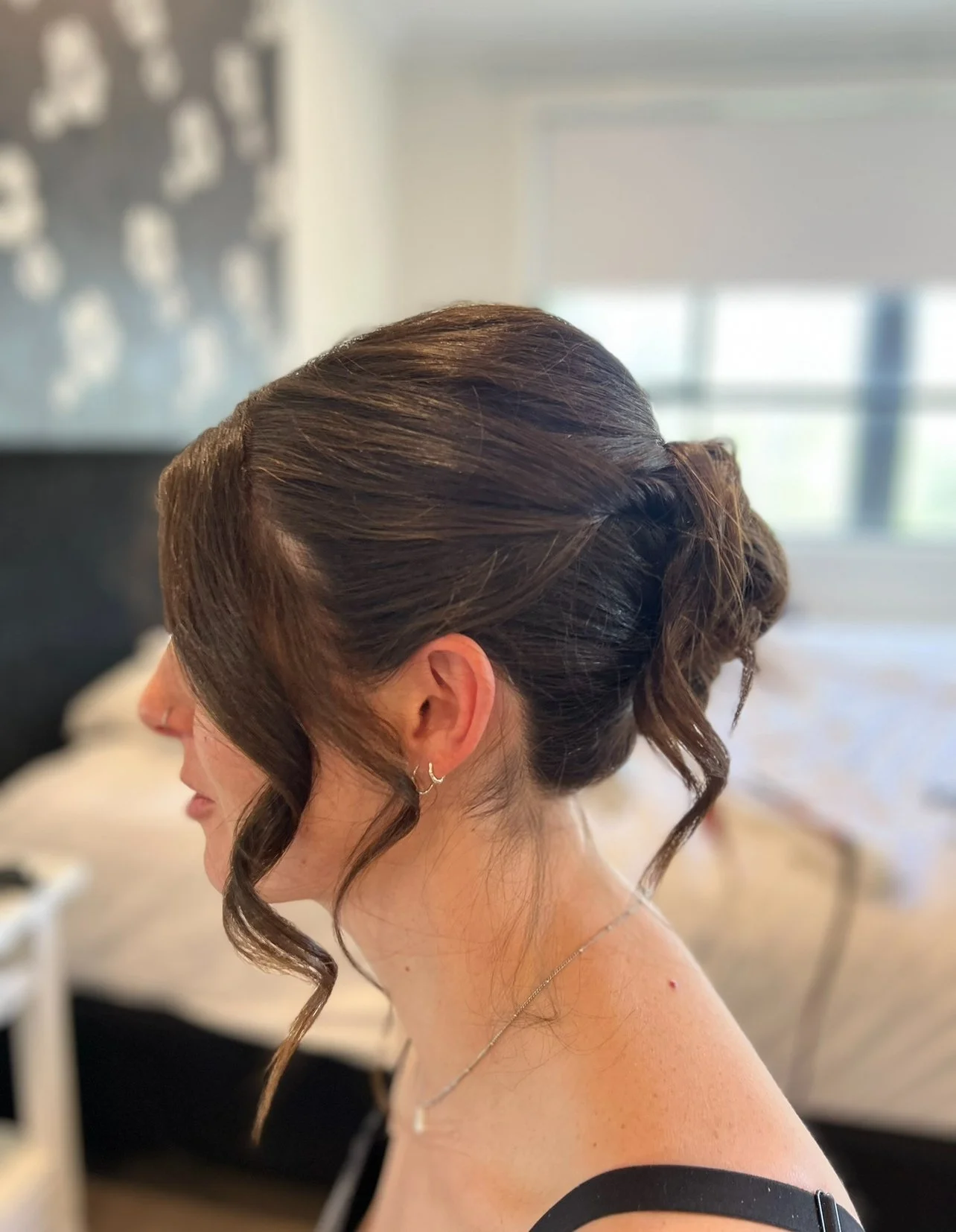 Close-up side view of a woman's elegant updo hairstyle with loose curls, earrings, and necklace in a softly lit bedroom.
