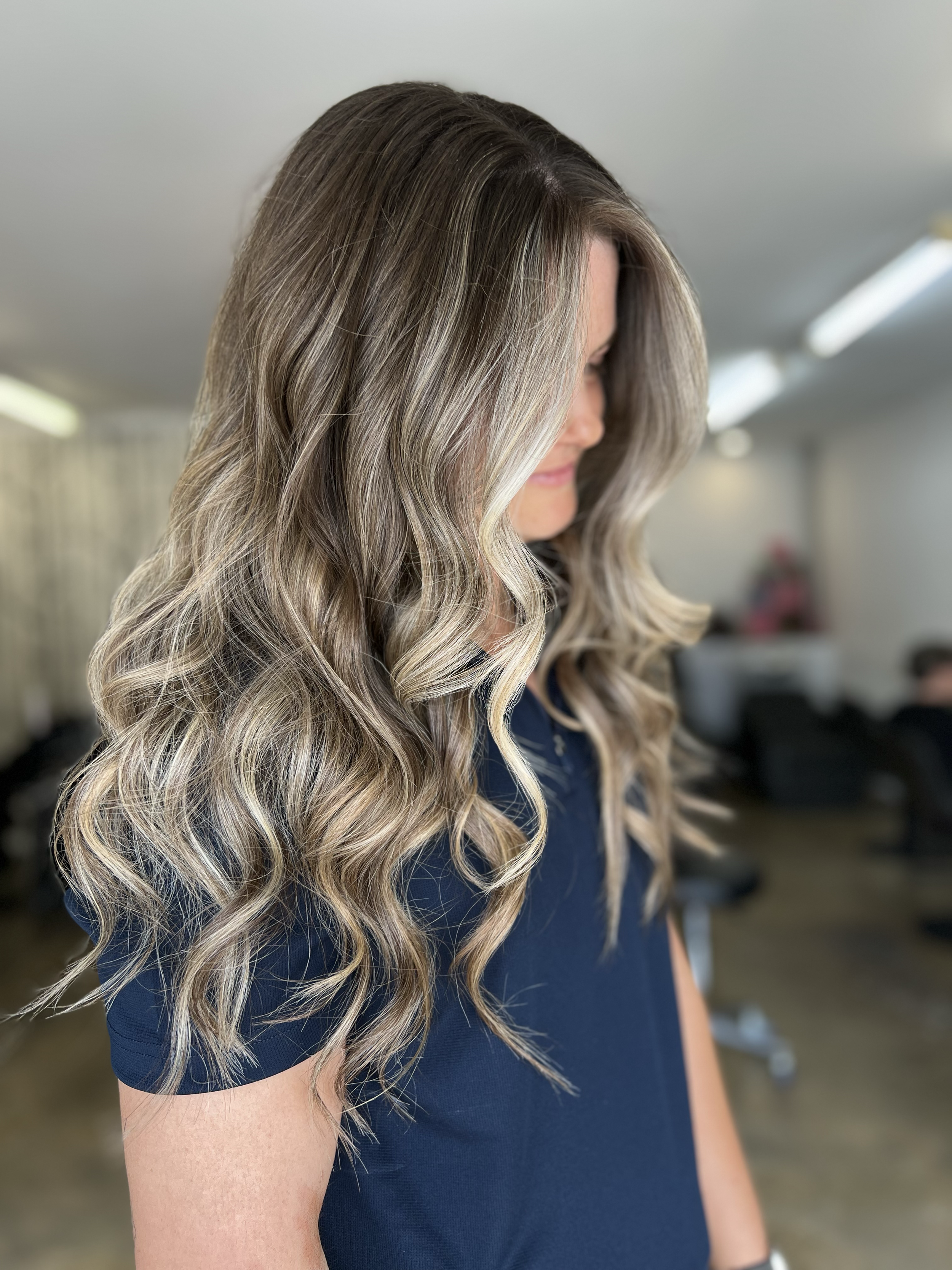 Woman with long, wavy blonde hair wearing a navy blue top in salon.