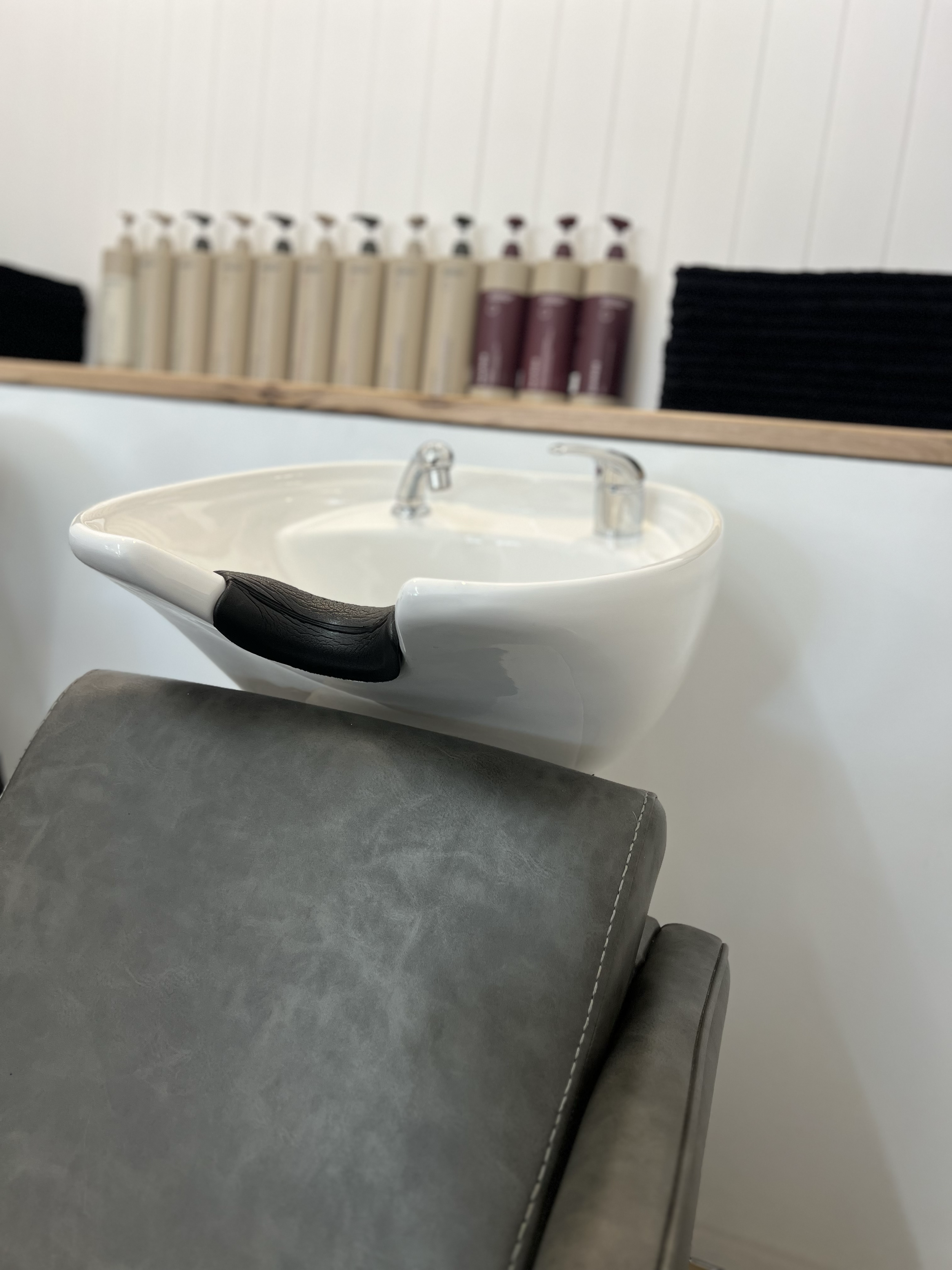 Close-up of a white shampoo bowl with black cushioned headrest, positioned above a grey salon chair, with shelves of hair products in bottles in the background.