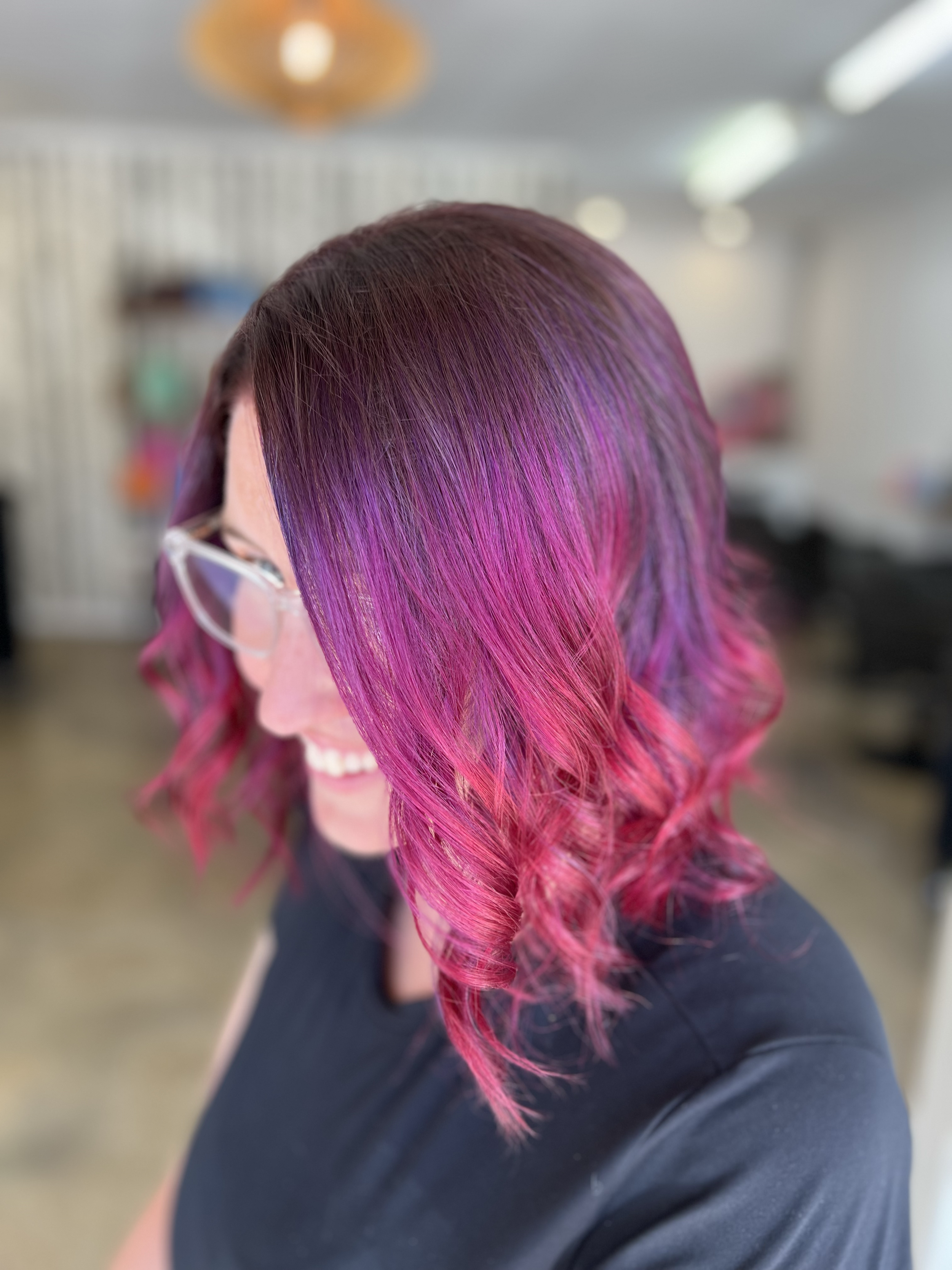A woman with shoulder-length hair styled in loose waves, colored with a gradient from dark purple at the roots to bright pink at the tips, wearing glasses and a black top, smiling while looking down.