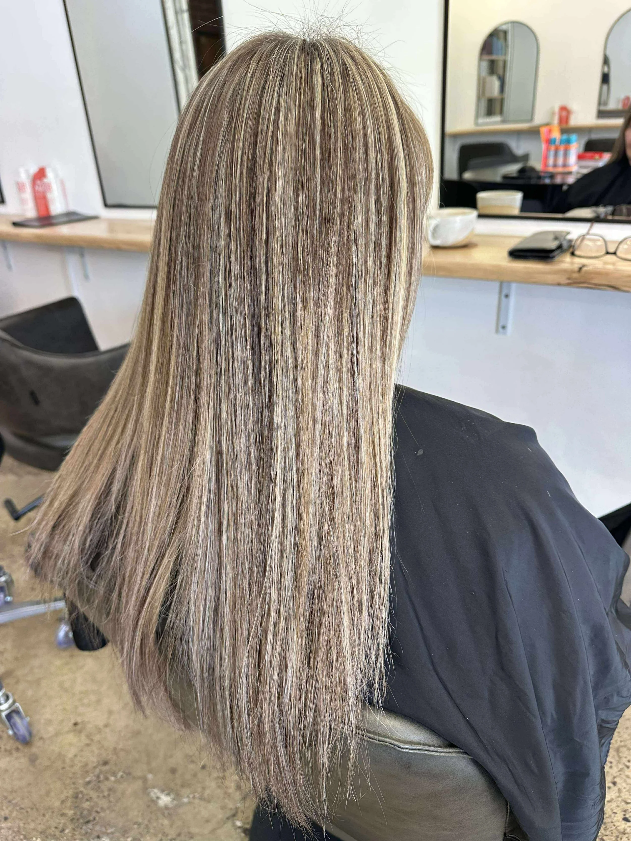 Back view of a woman with long, straight blonde hair sitting in a salon chair, with styling tools and mirror reflections visible in the background. keratin treatment