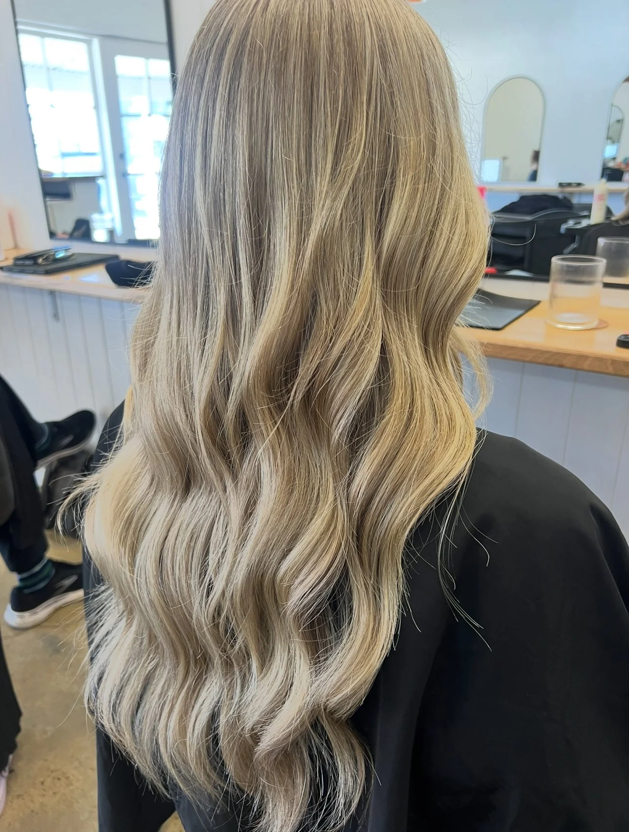 A woman with long, wavy blonde hair sitting in a salon chair with a black cape. The salon has large windows, mirrors, and various hair styling tools on a counter.