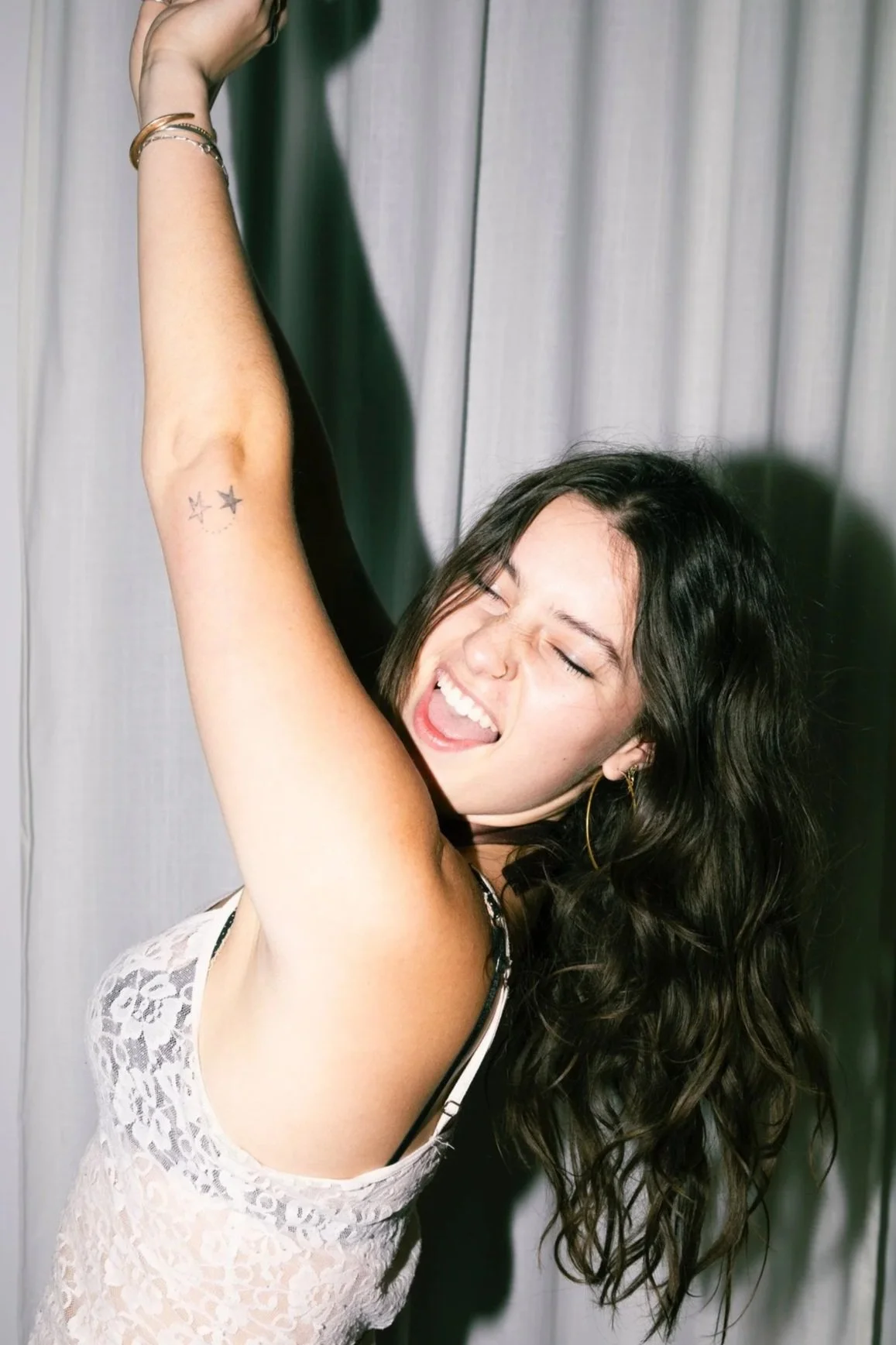 Young woman with dark wavy hair, wearing a white lace top, laughing with eyes closed, raising her left arm, standing in front of gray curtains.