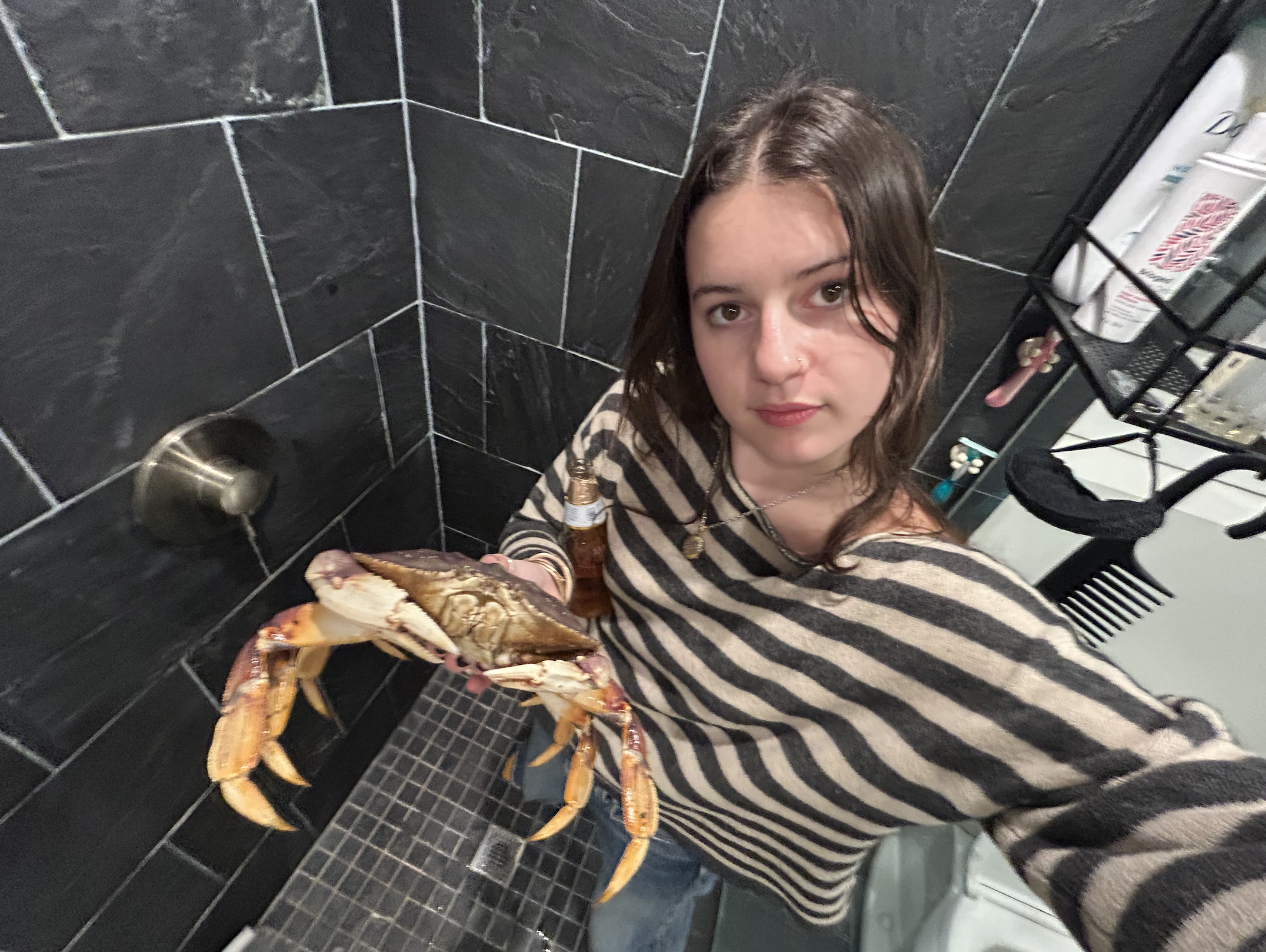 A young woman with brown hair and a striped shirt holding a large lobster in front of her at a black-tiled wall with a metal electric outlet and a shelf holding medical supplies.