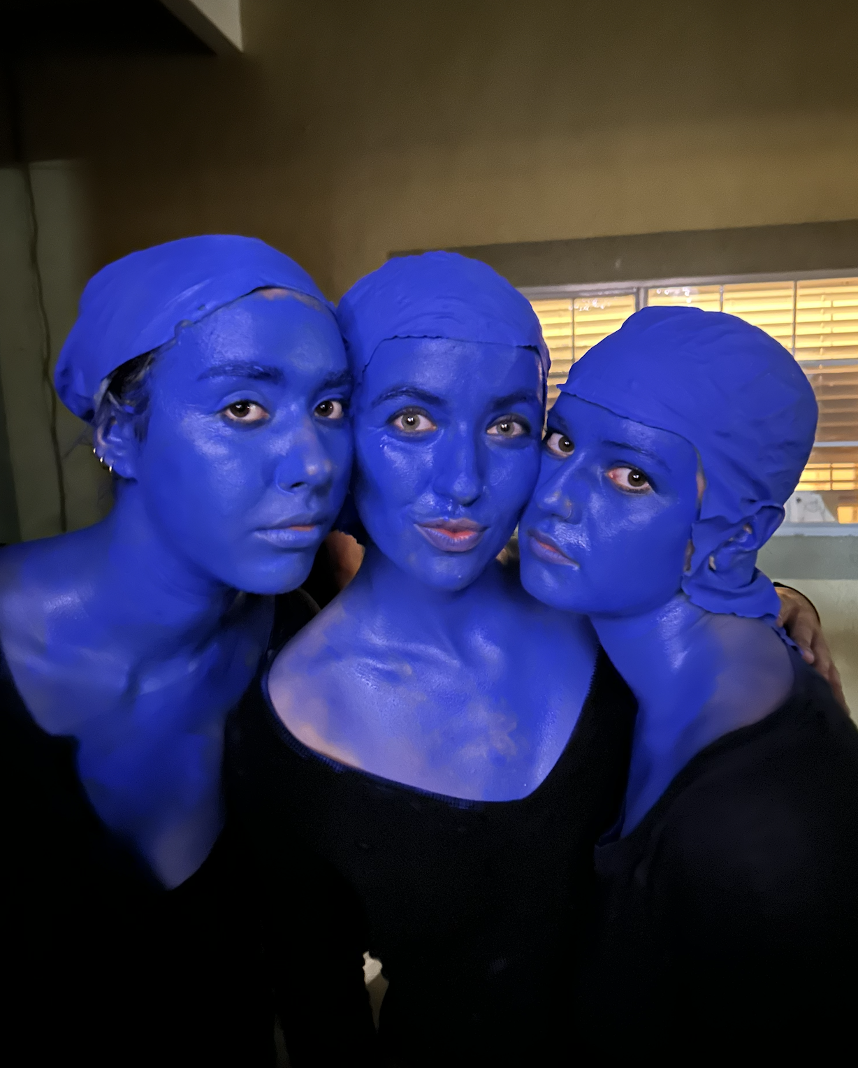 Three women with their faces and bodies painted entirely in blue, wearing matching blue head coverings, taking a selfie together indoors near a window with blinds.
