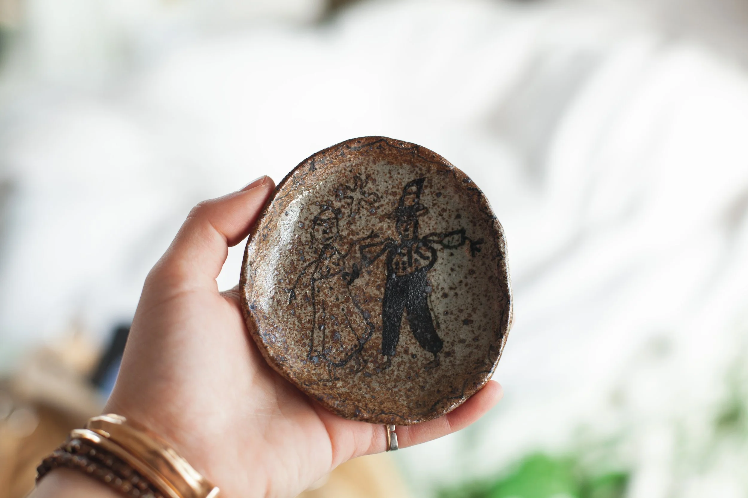 Person holding a ceramic plate with a black figure and abstract designs, against a blurred background.