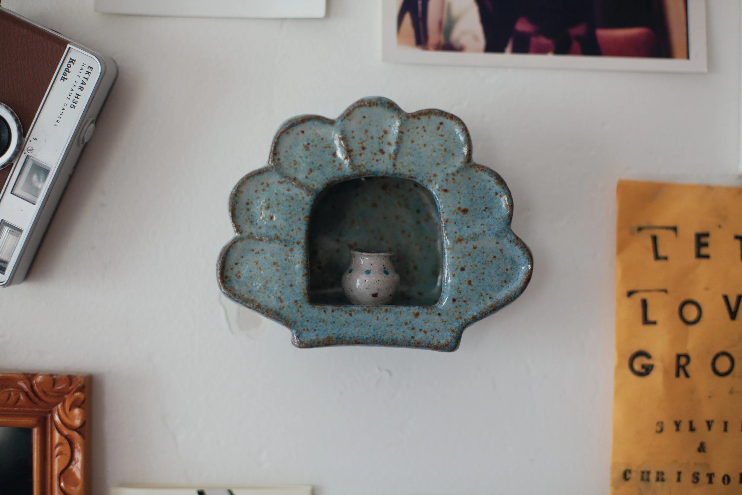A ceramic flower-shaped holder with blue glaze and brown speckles, positioned on a white surface alongside other decorative objects and photographs.