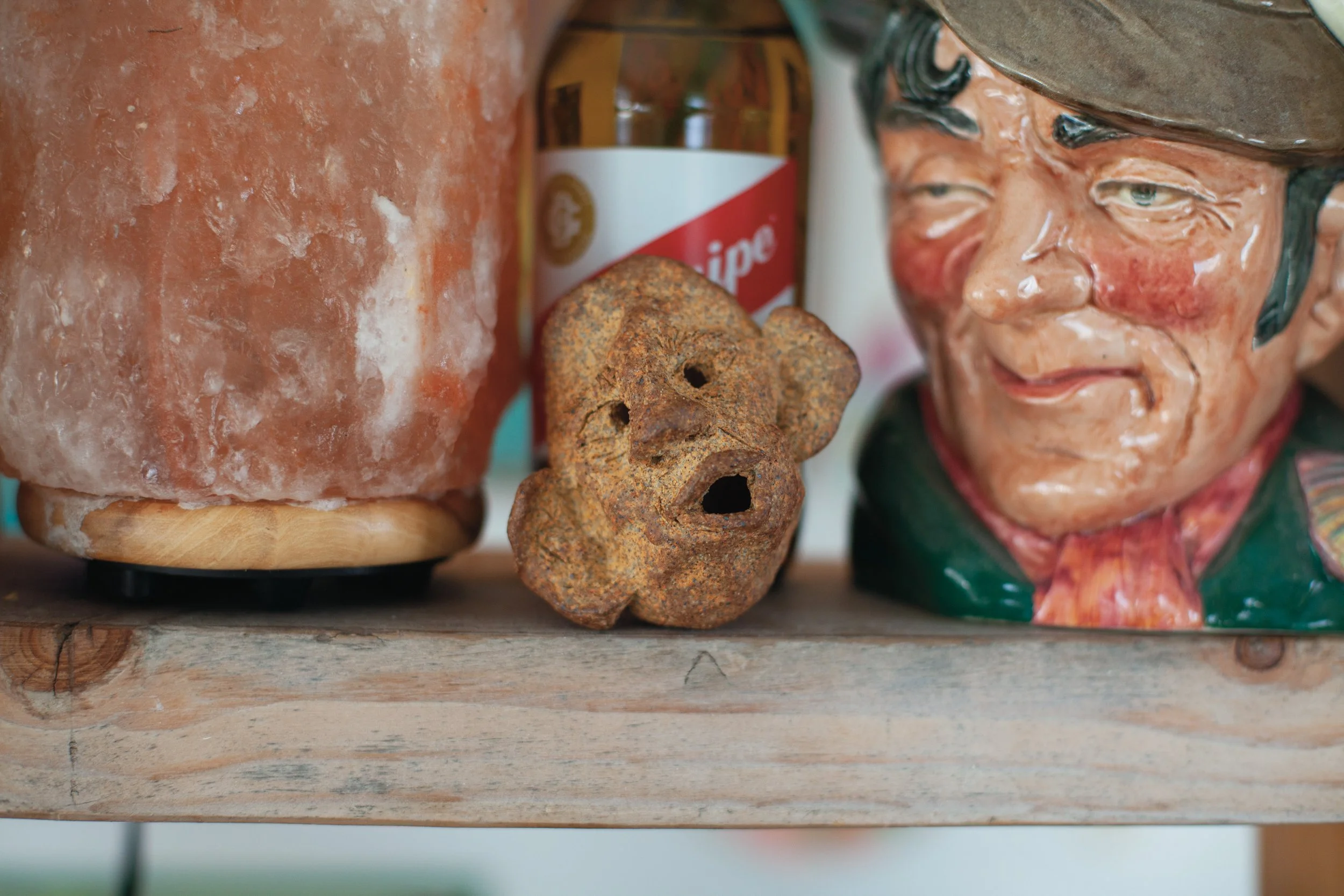 Close-up of salt lamp, rusted flower-shaped object, ceramic mug with face, and salt shaker on a wooden shelf.