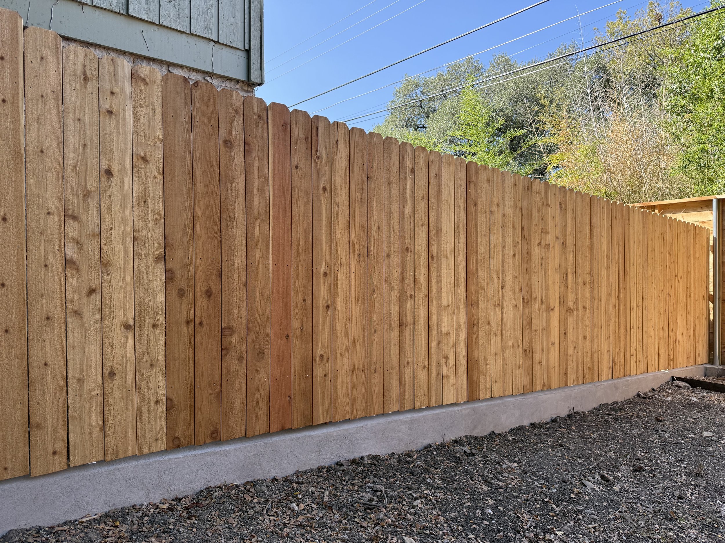 New wooden fence installed along the perimeter of a yard, with a concrete base and dirt ground in front, under a clear blue sky.