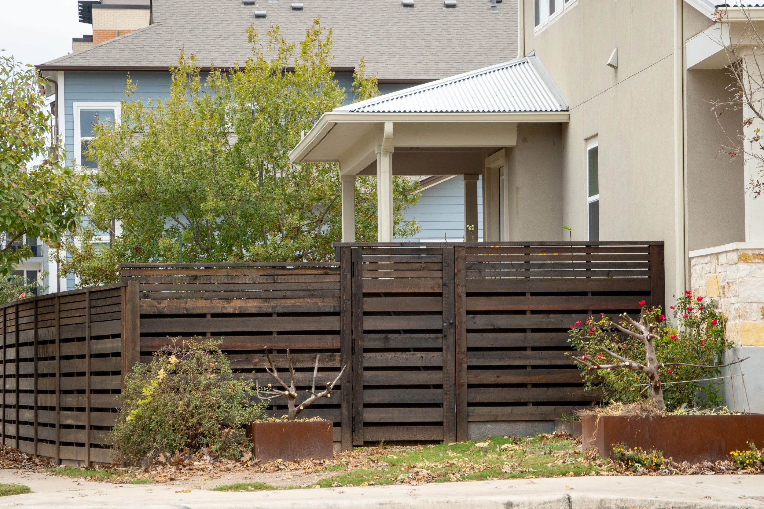 Residential horizontal shadow box privacy fence, small trees, and a house with a covered porch and metal roof.