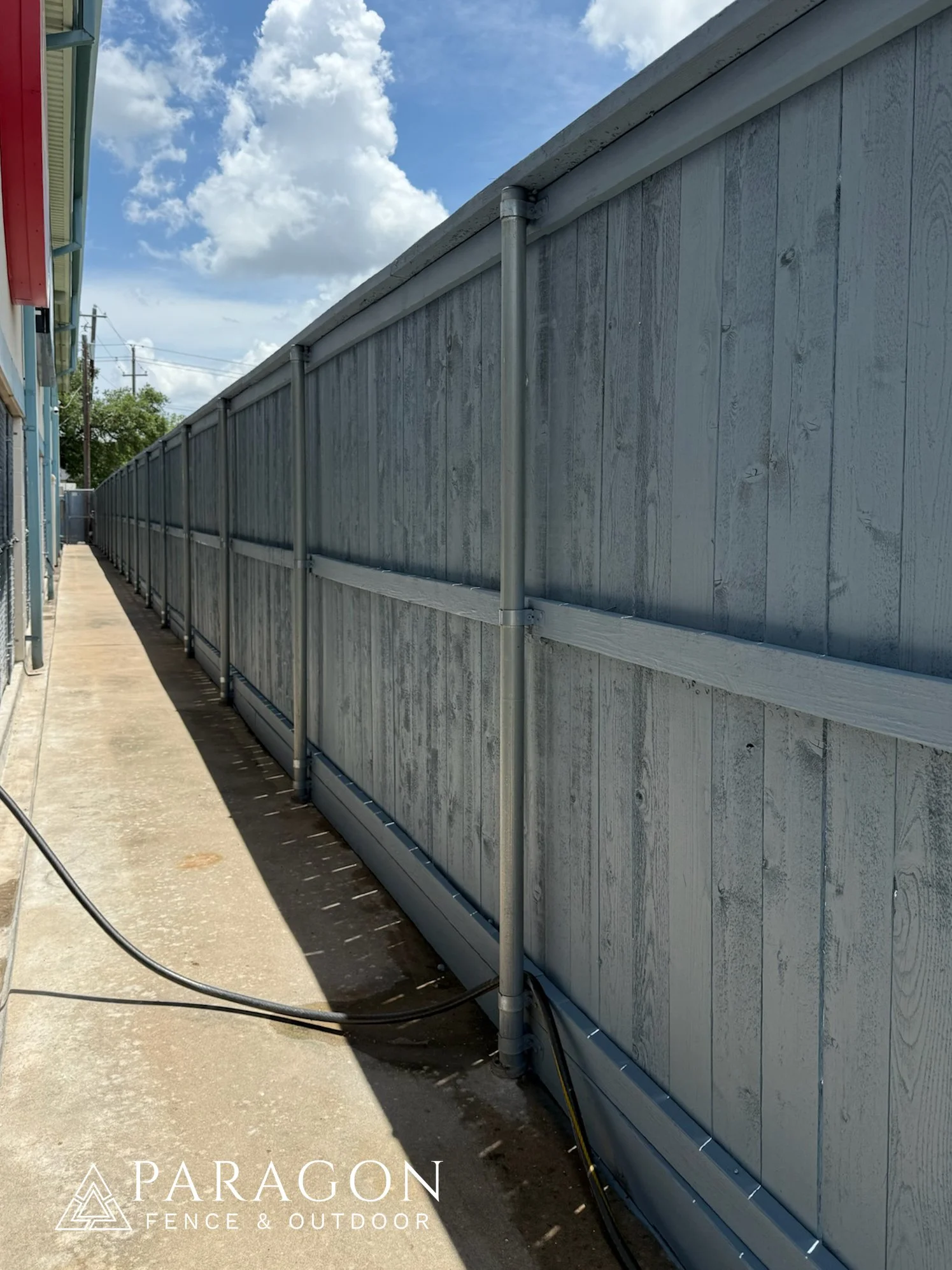 Gray wooden privacy fence with metal posts along a concrete sidewalk, with an electrical cable on the ground and a blue sky with clouds in the background.