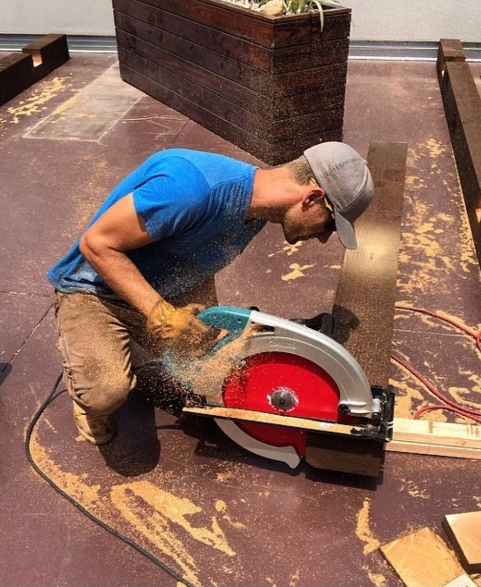 A man wearing a gray cap, glasses, and a blue t-shirt is cutting a wooden board with a red and silver circular saw. Sawdust is flying around as he works on a woodworking project.