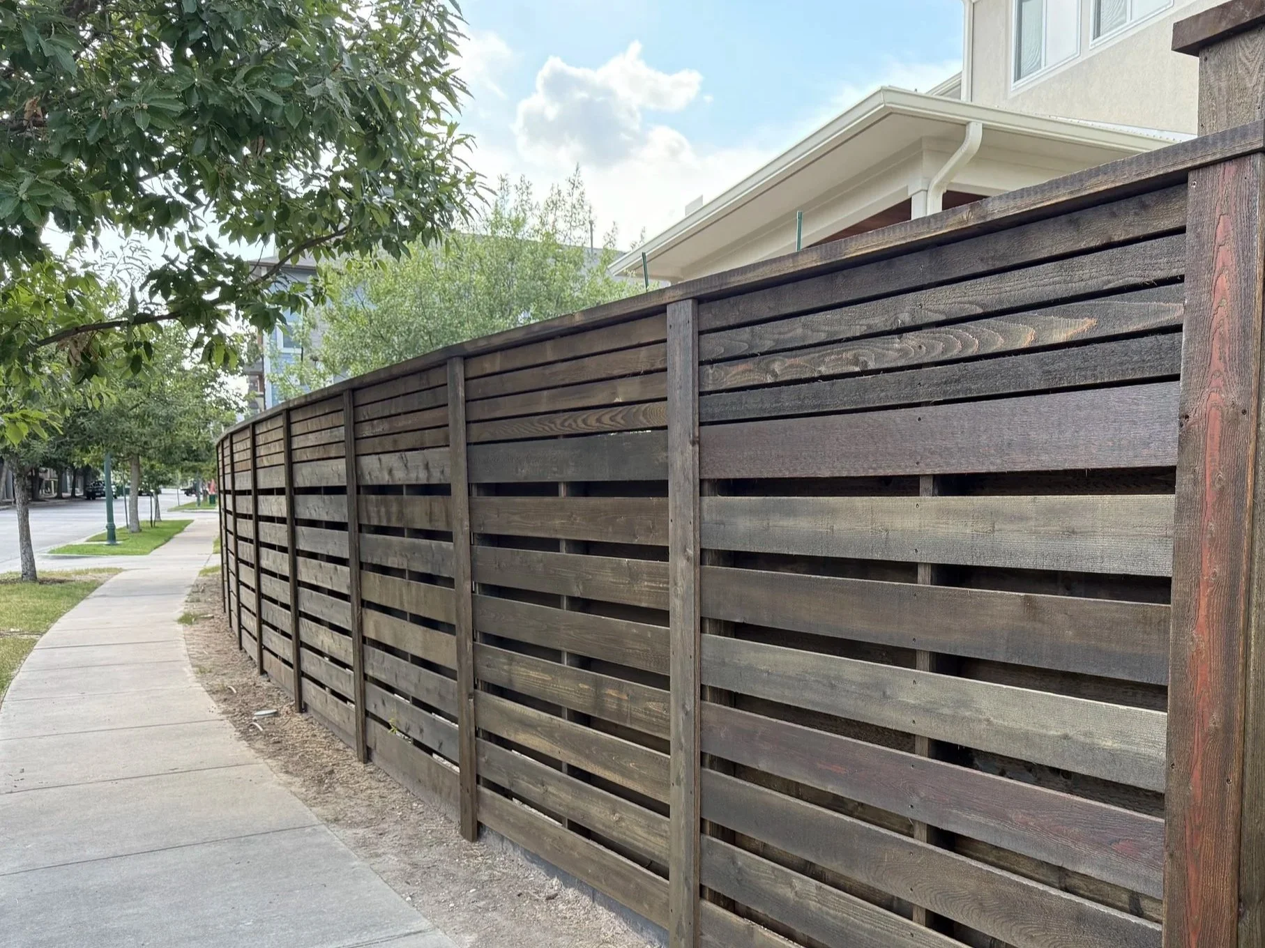 Wooden privacy fence along a sidewalk with trees in the background and a residential building.
