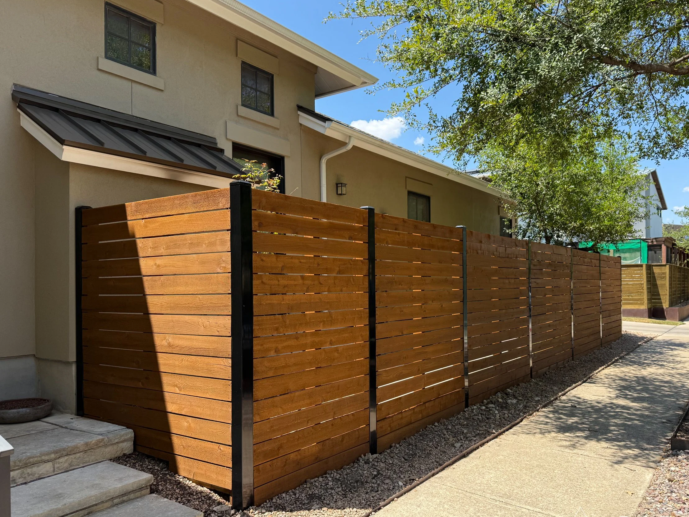 A wooden privacy fence along a sidewalk in a residential neighborhood with trees and houses.
