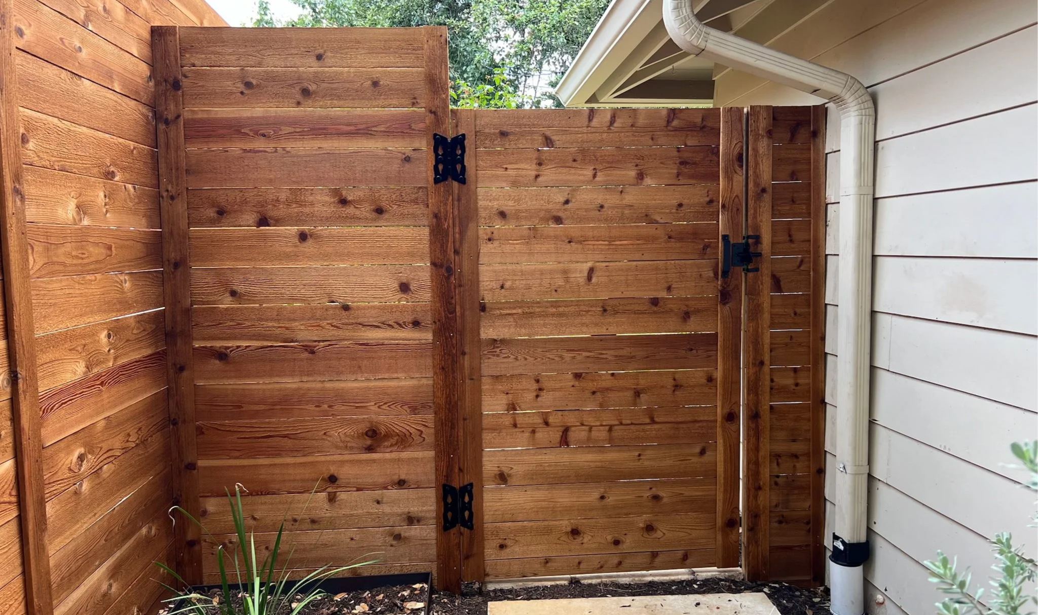 Wooden outdoor privacy fence with gate, next to house wall with downspout, planting bed with green plant in foreground.